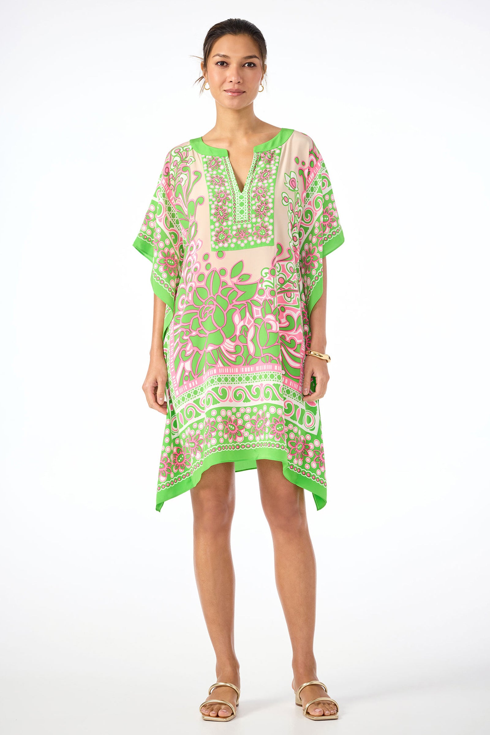 A woman wears the Theodora Caftan Dress in Petra Floral silk, a loose-fitting, knee-length dress with pink and green patterns, styled with bracelets and sandals against a white background.