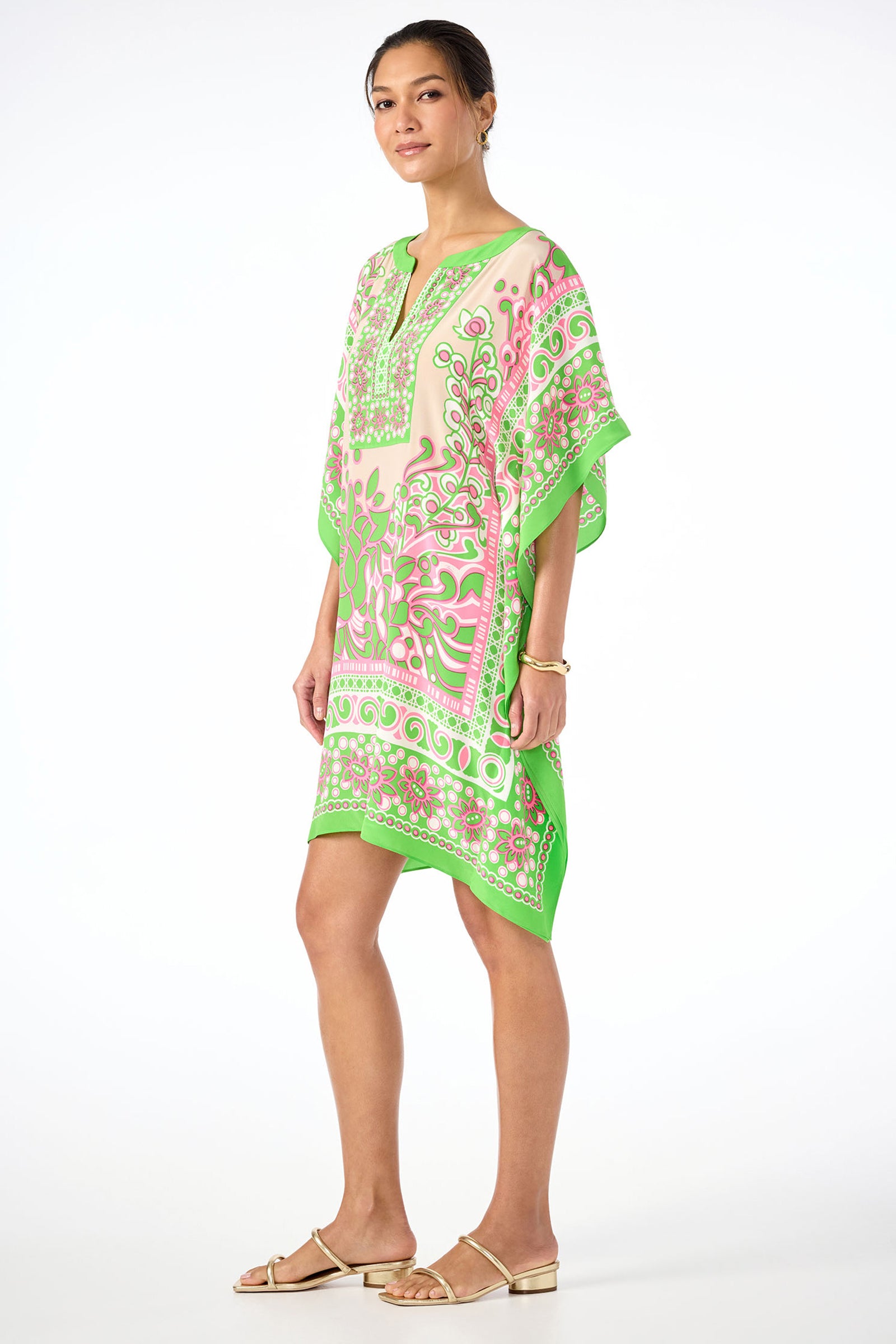 A woman stands sideways against a white background, wearing Theodora Caftan Dress - Petra Floral Silk with green, pink, and white patterns. Her hair is up, she wears minimal jewelry and gold flat sandals.