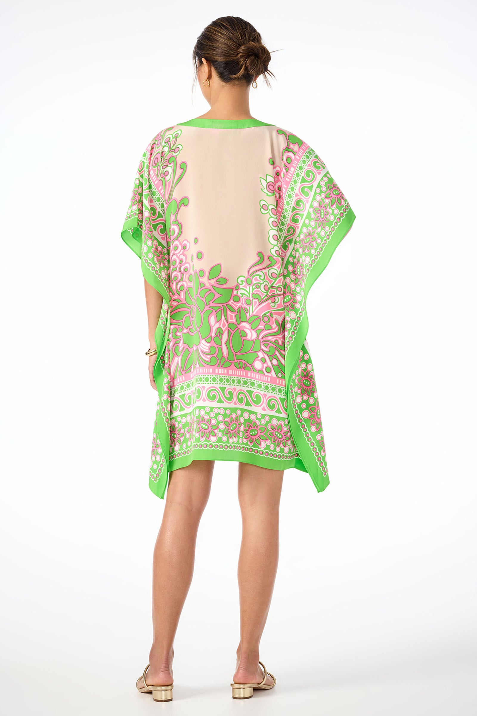 A woman stands barefoot with her back to the camera, hair in a bun, wearing Theodora Caftan Dress - Petra Floral Silk, a loose knee-length kimono-style dress featuring a colorful pink, green, and beige floral pattern.