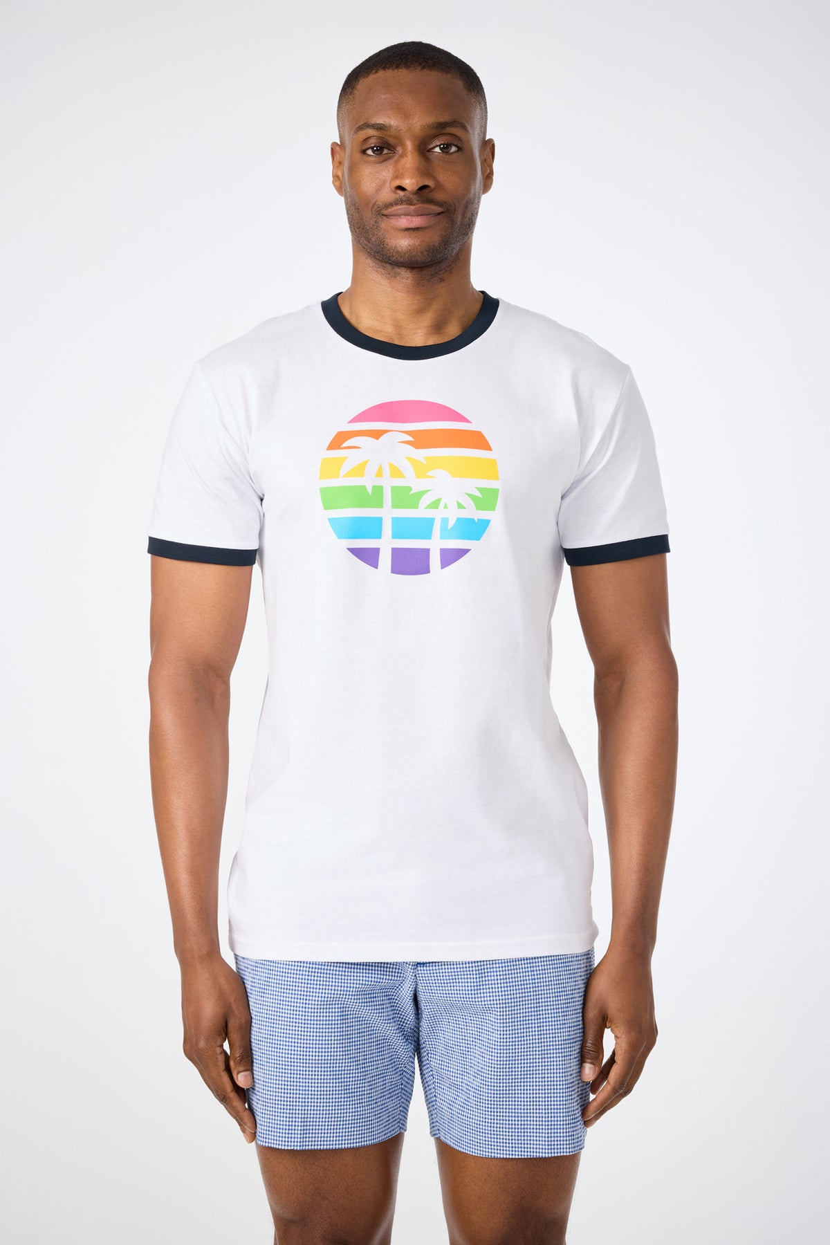 A man faces forward against a white background, wearing the Short-Sleeve Crewneck T-Shirt - Pride in WHITE with a rainbow-striped circle and palm trees graphic, paired with blue checkered shorts.