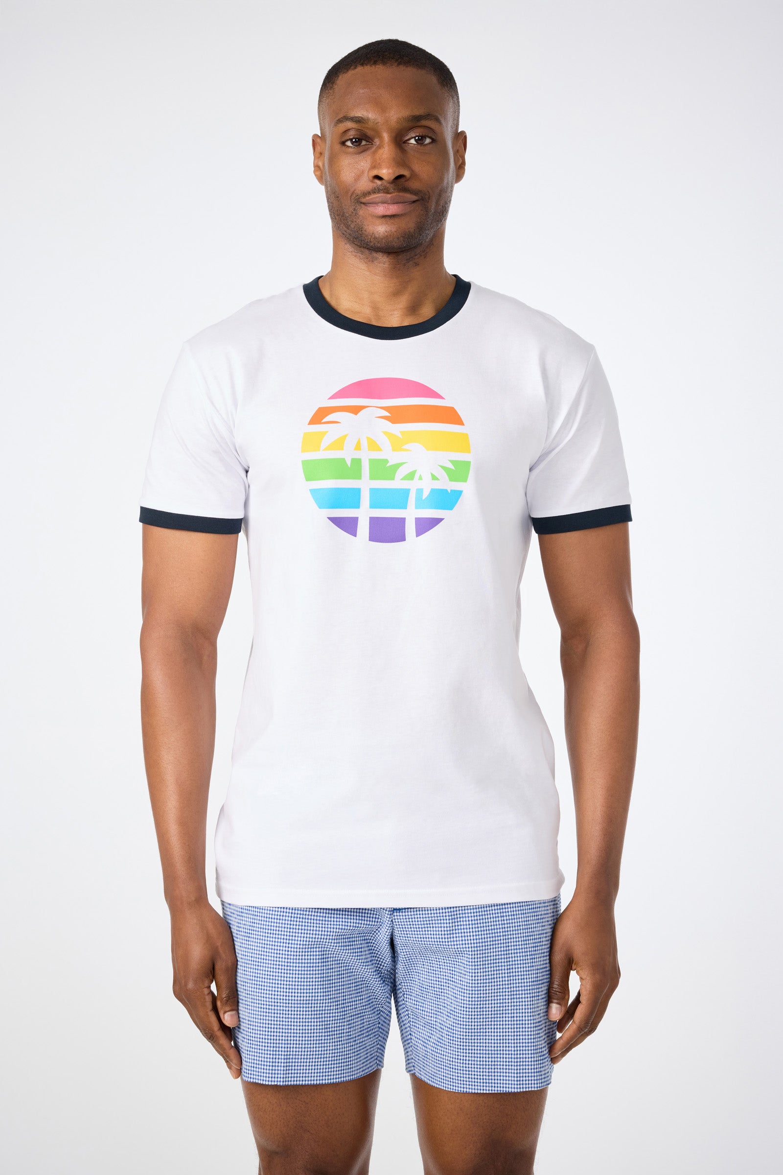 A man faces forward against a white background, wearing the Short-Sleeve Crewneck T-Shirt - Pride in WHITE with a rainbow-striped circle and palm trees graphic, paired with blue checkered shorts.