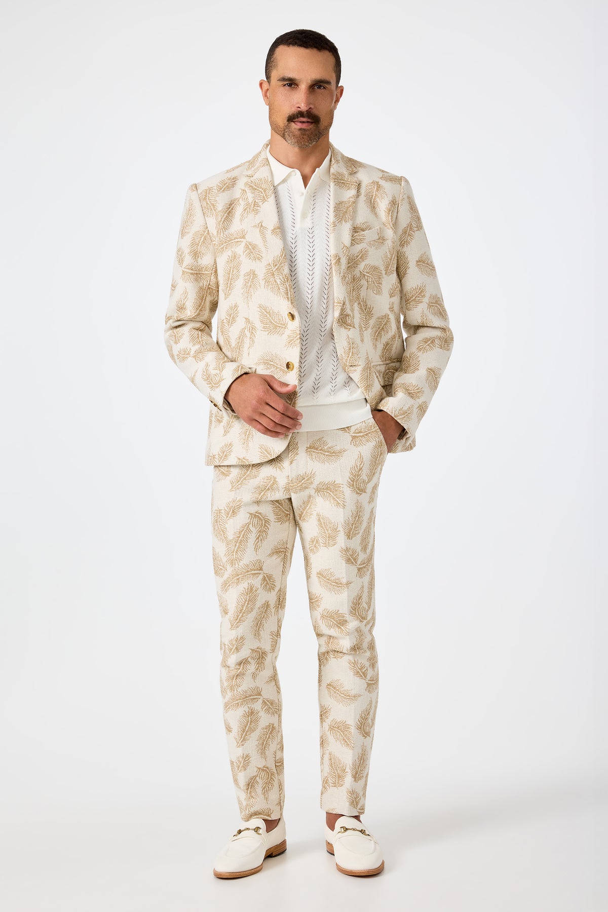A man with a mustache wears the Thurston Blazer in Desert Fronds Jacquard over a white shirt and loafers, standing against a plain white background.