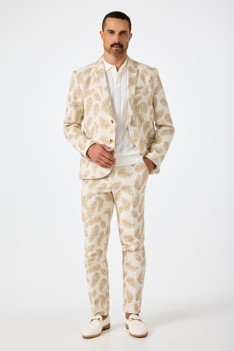 A man with a mustache wears the Thurston Blazer in Desert Fronds Jacquard over a white shirt and loafers, standing against a plain white background.