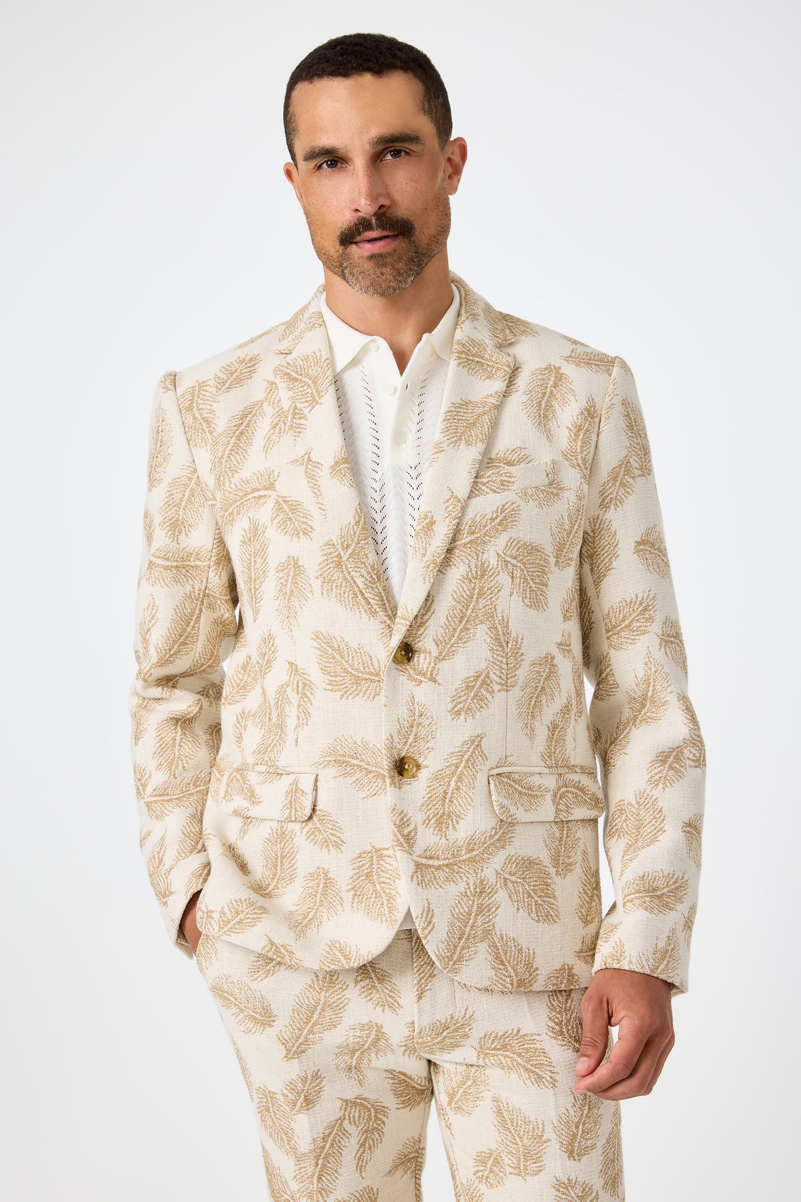 A man with short dark hair and a mustache wears the Thurston Blazer in Desert Fronds Jacquard over a white shirt, with one hand in his pocket, standing against a plain white background.
