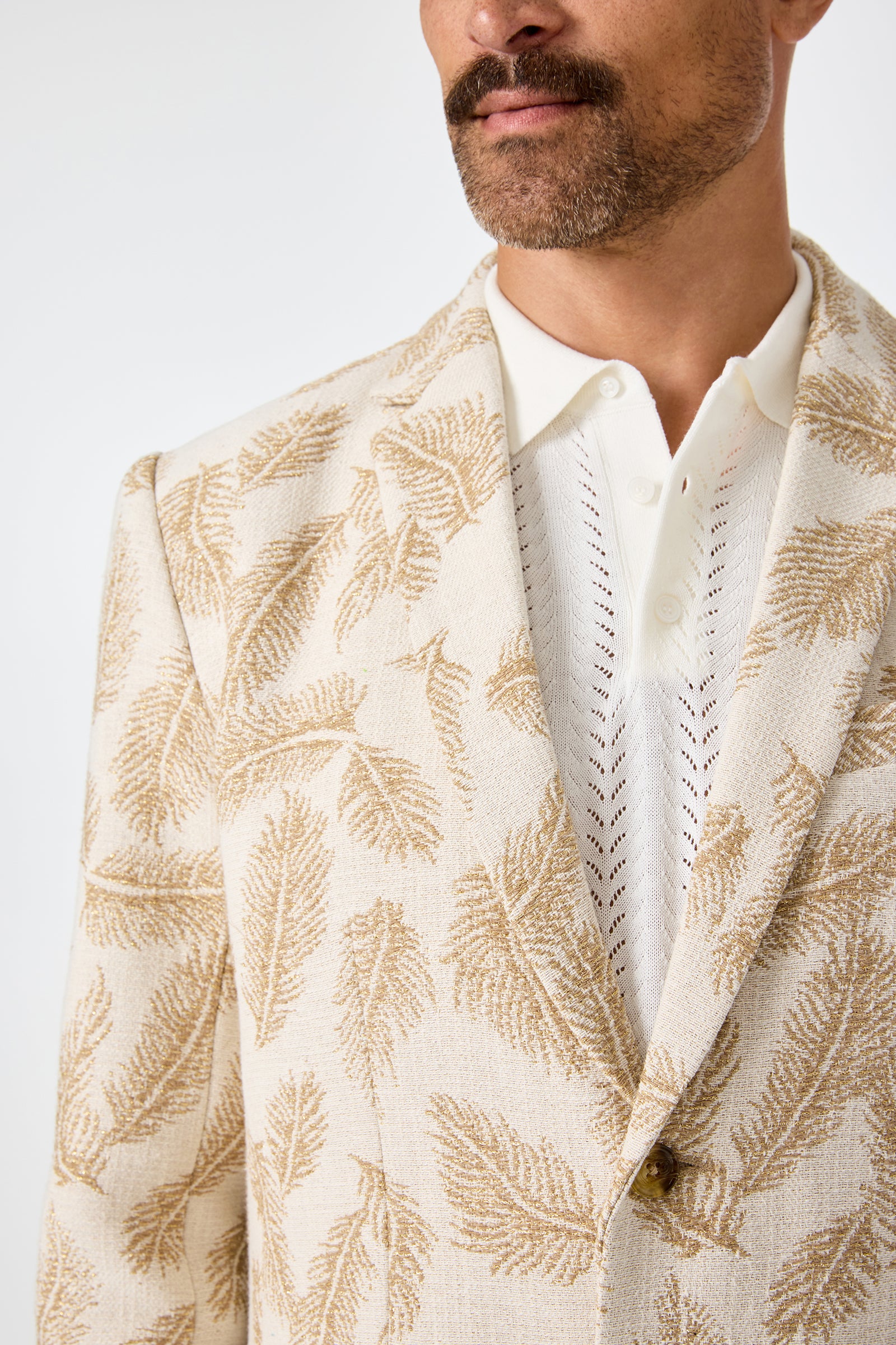 A man with a mustache wears the Thurston Blazer in Desert Fronds Jacquard—a cream blazer with a gold leaf pattern—over a white perforated collared shirt. Only his lower face and upper torso are visible.