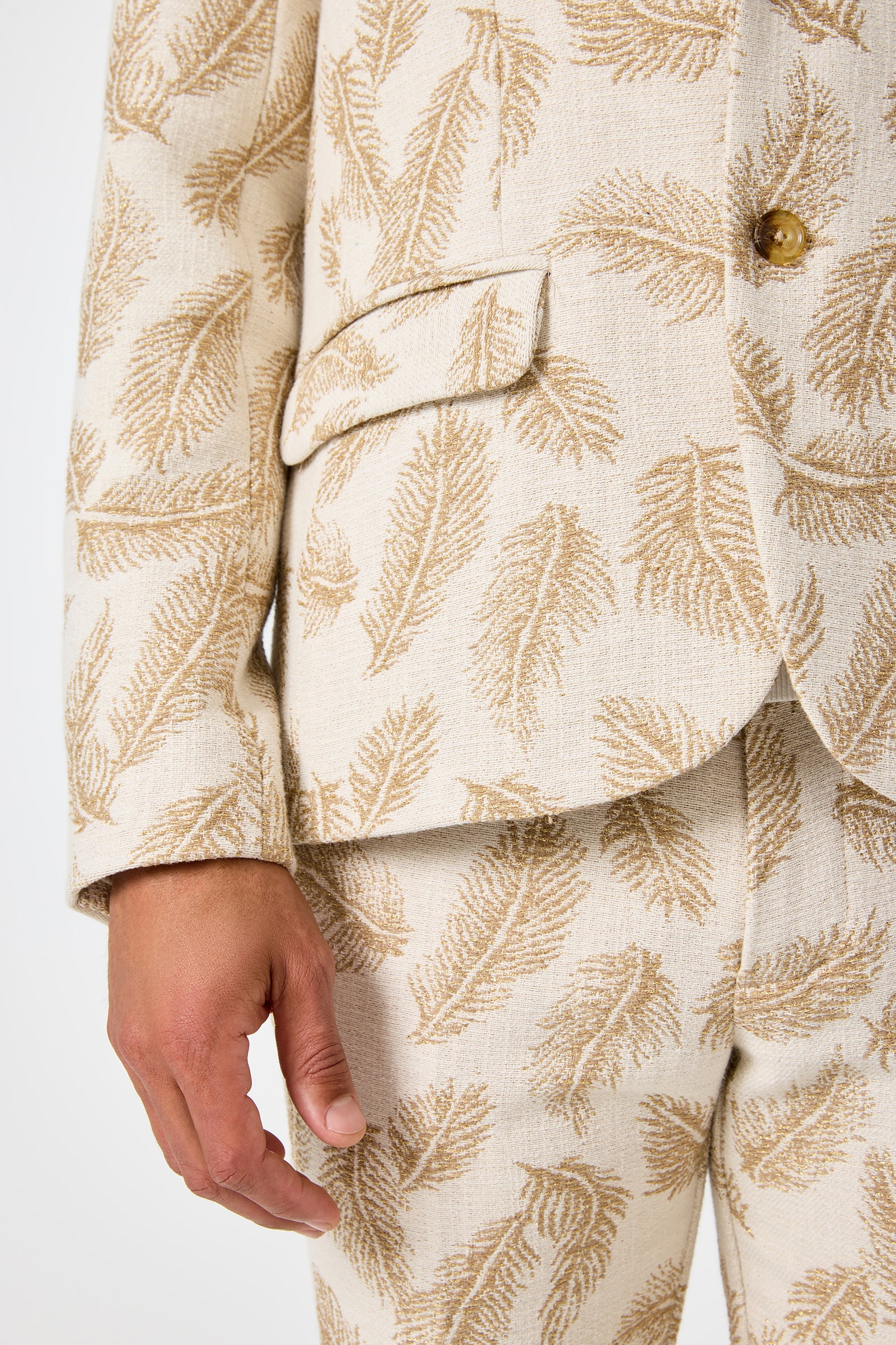 A close-up of the Thurston Blazer in Desert Fronds Jacquard shows the lower half of the cream-colored jacket with a gold leaf pattern, matching pants, and the persons left hand at their side.