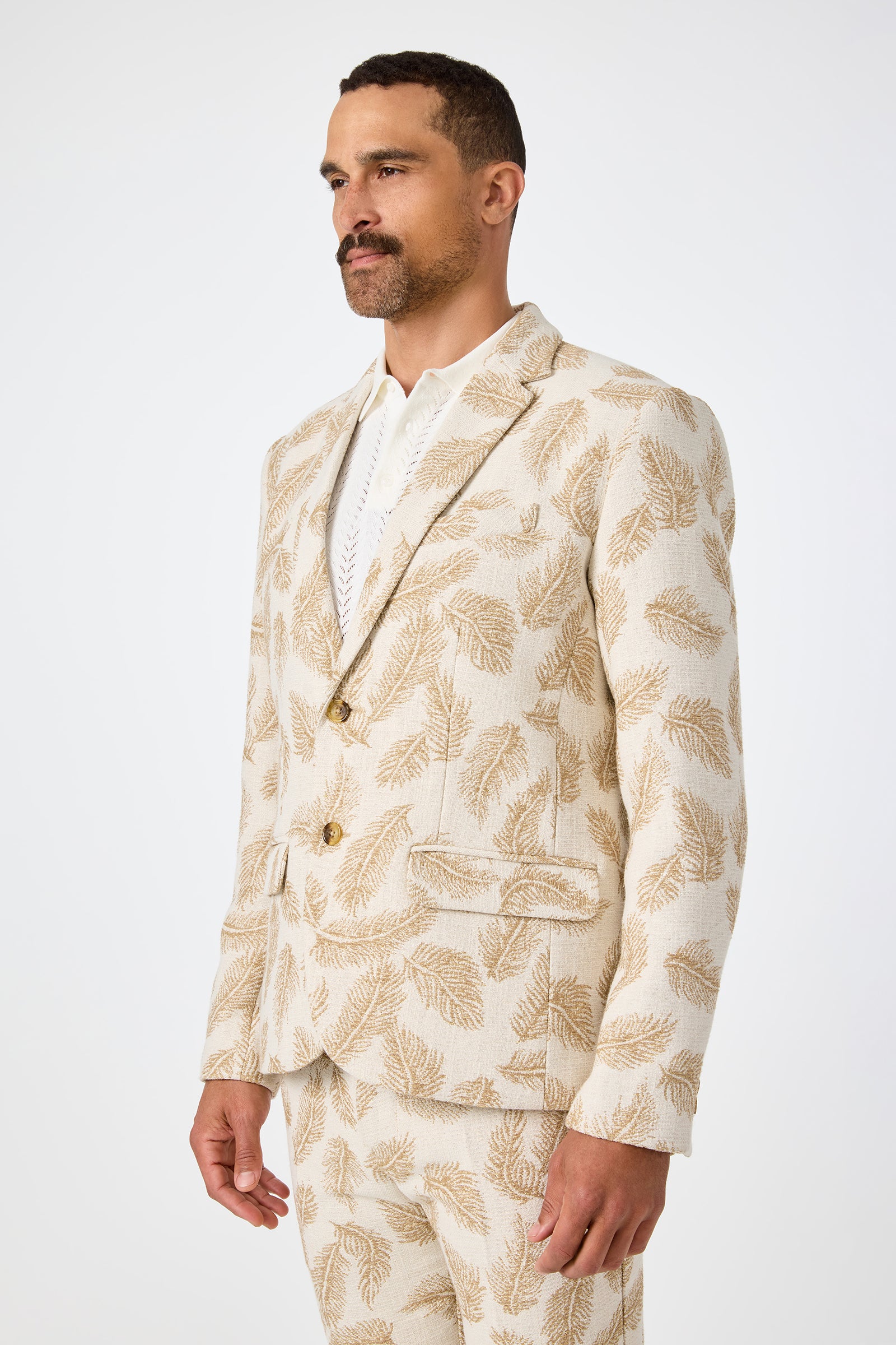 A man models the Thurston Blazer in Desert Fronds Jacquard—a beige suit with a brown leaf design—over a white shirt, standing against a plain white background and gazing slightly to the side.