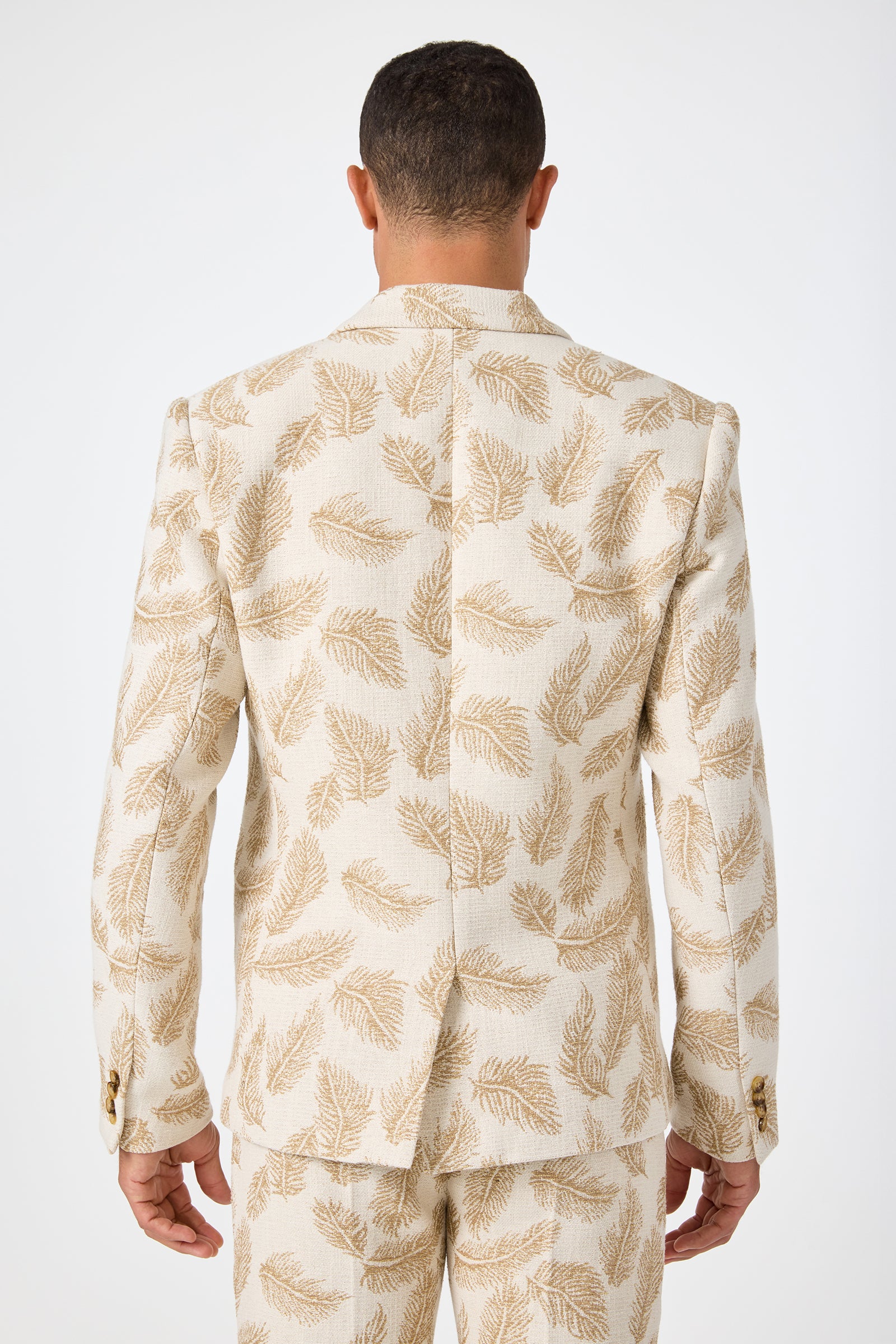 A man faces away from the camera, wearing the Thurston Blazer in Desert Fronds Jacquard—a cream suit with a light brown leaf pattern on both jacket and trousers—against a plain white background.