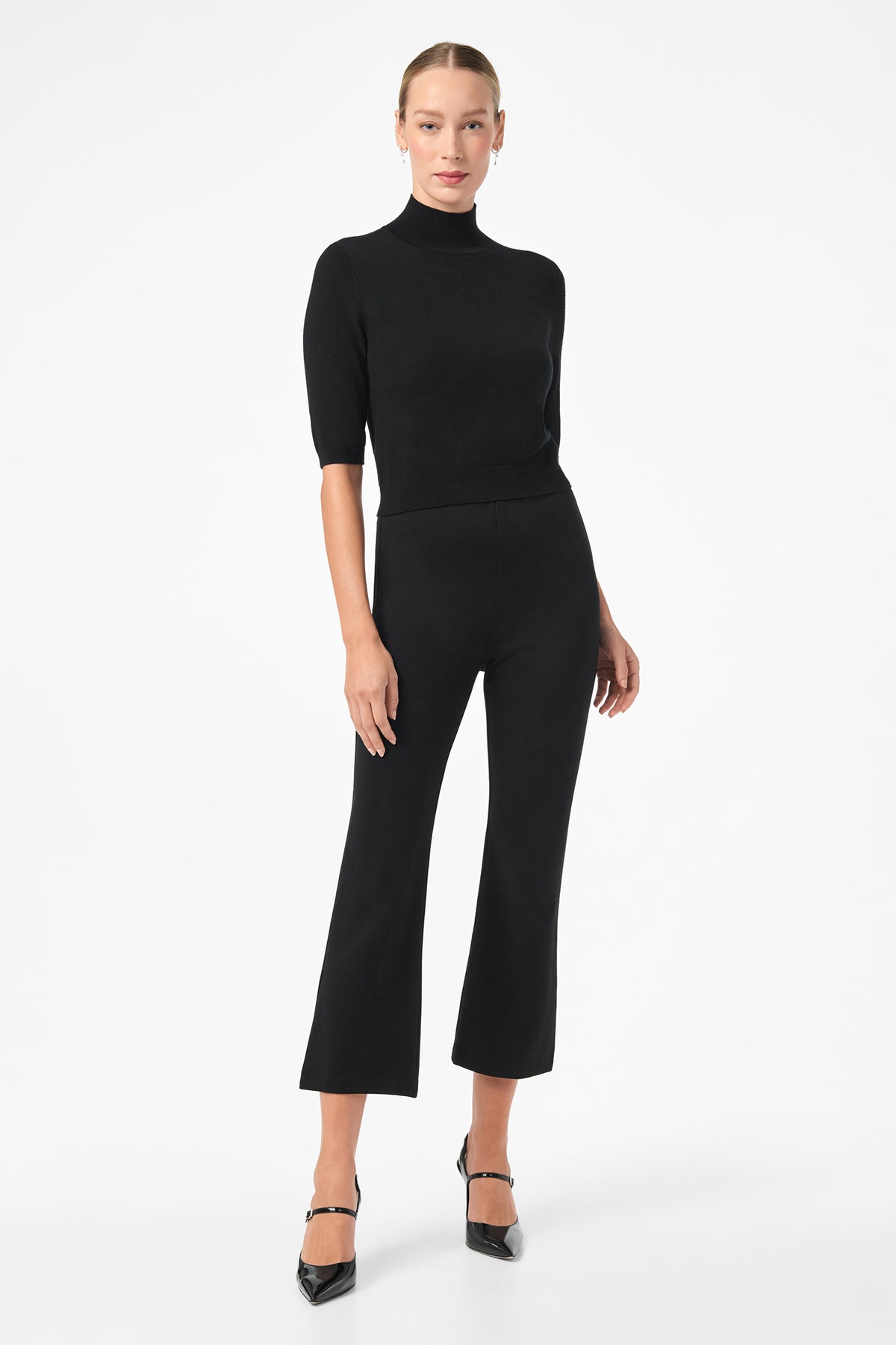 A woman stands against a white background wearing black Sorrel Kick-Flare Pants, a fitted black turtleneck top, and black heels. Her hair is pulled back, and she looks at the camera with a neutral expression.