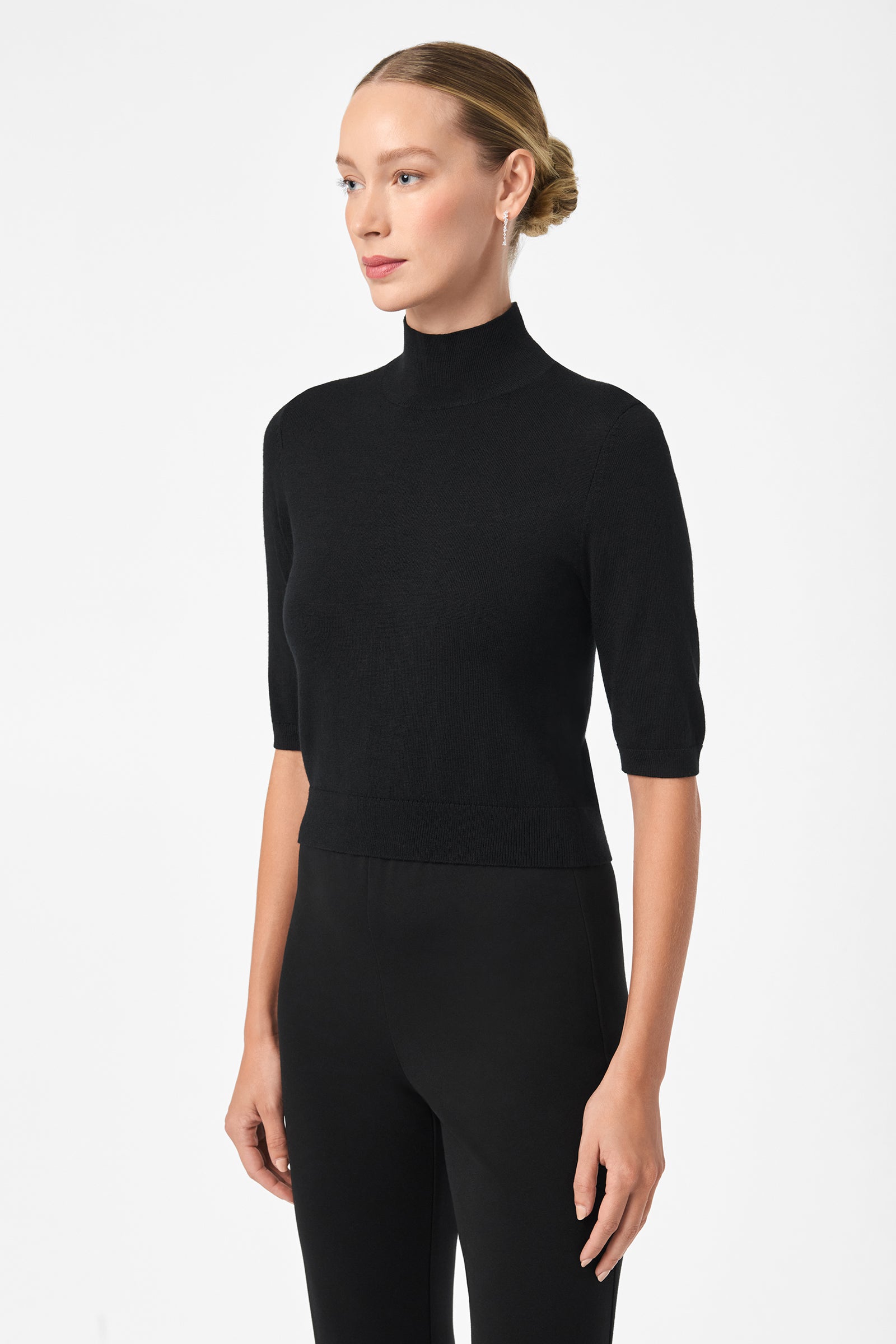 A woman with blonde hair in a neat bun wears the fitted black Mali Mock Neck top and black pants, standing against a white background and looking slightly to the side.