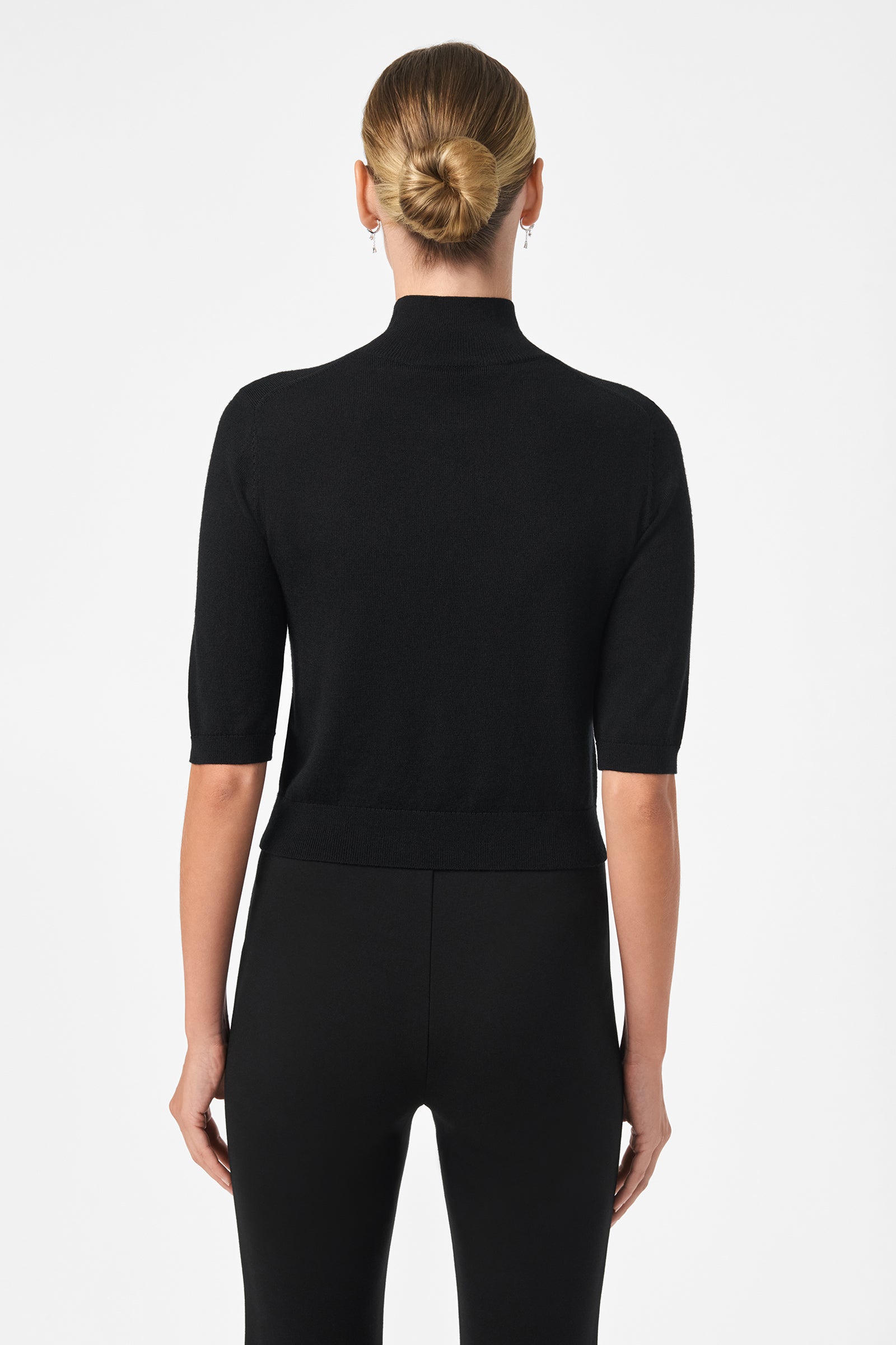 A woman with blonde hair in a sleek bun stands with her back to the camera, wearing the fitted black Mali Mock Neck short-sleeve top and black pants against a plain white background.