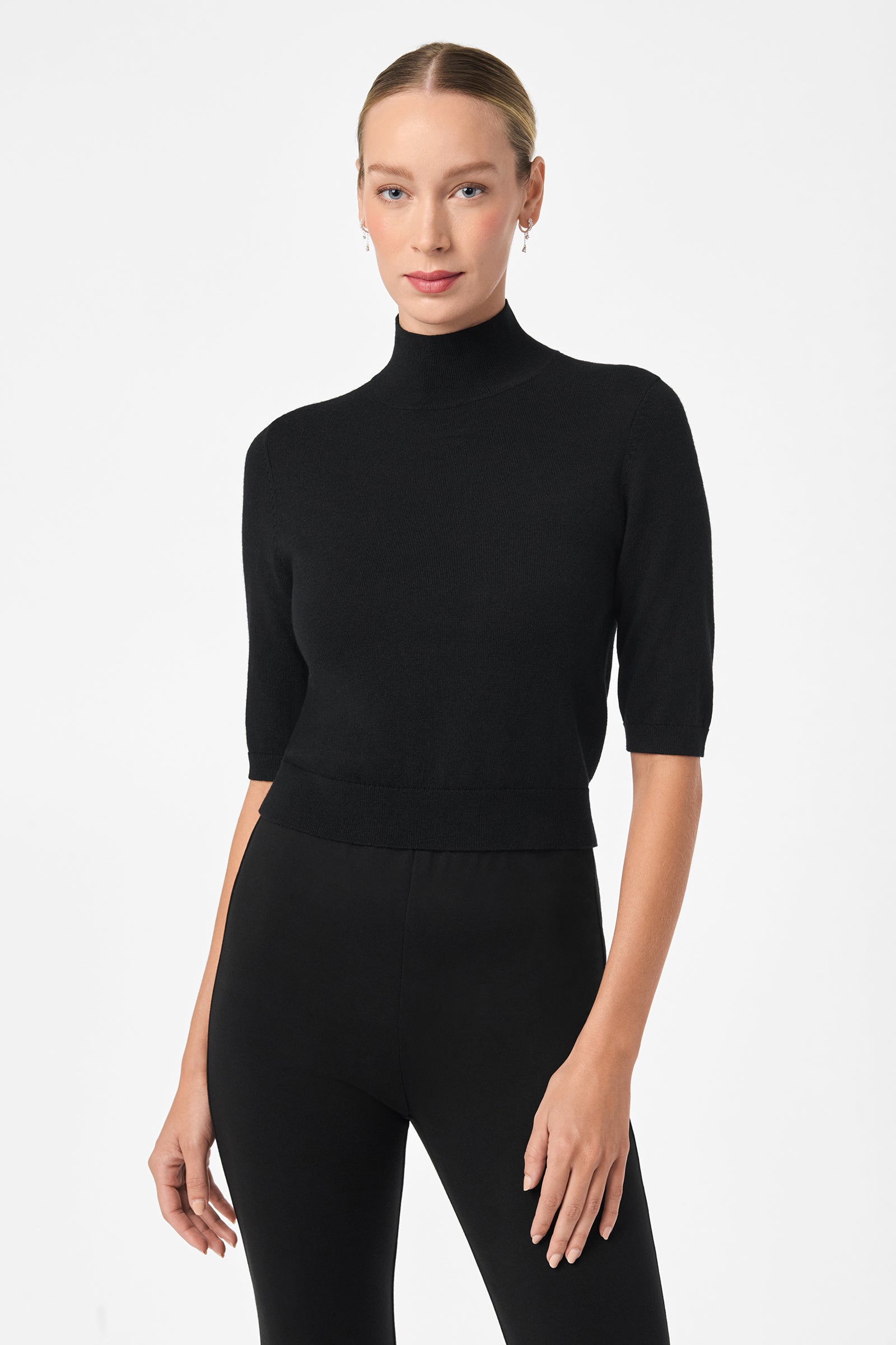A woman with blonde hair in a neat bun wears the fitted black Mali Mock Neck short-sleeve sweater and black pants, standing confidently against a plain white background.
