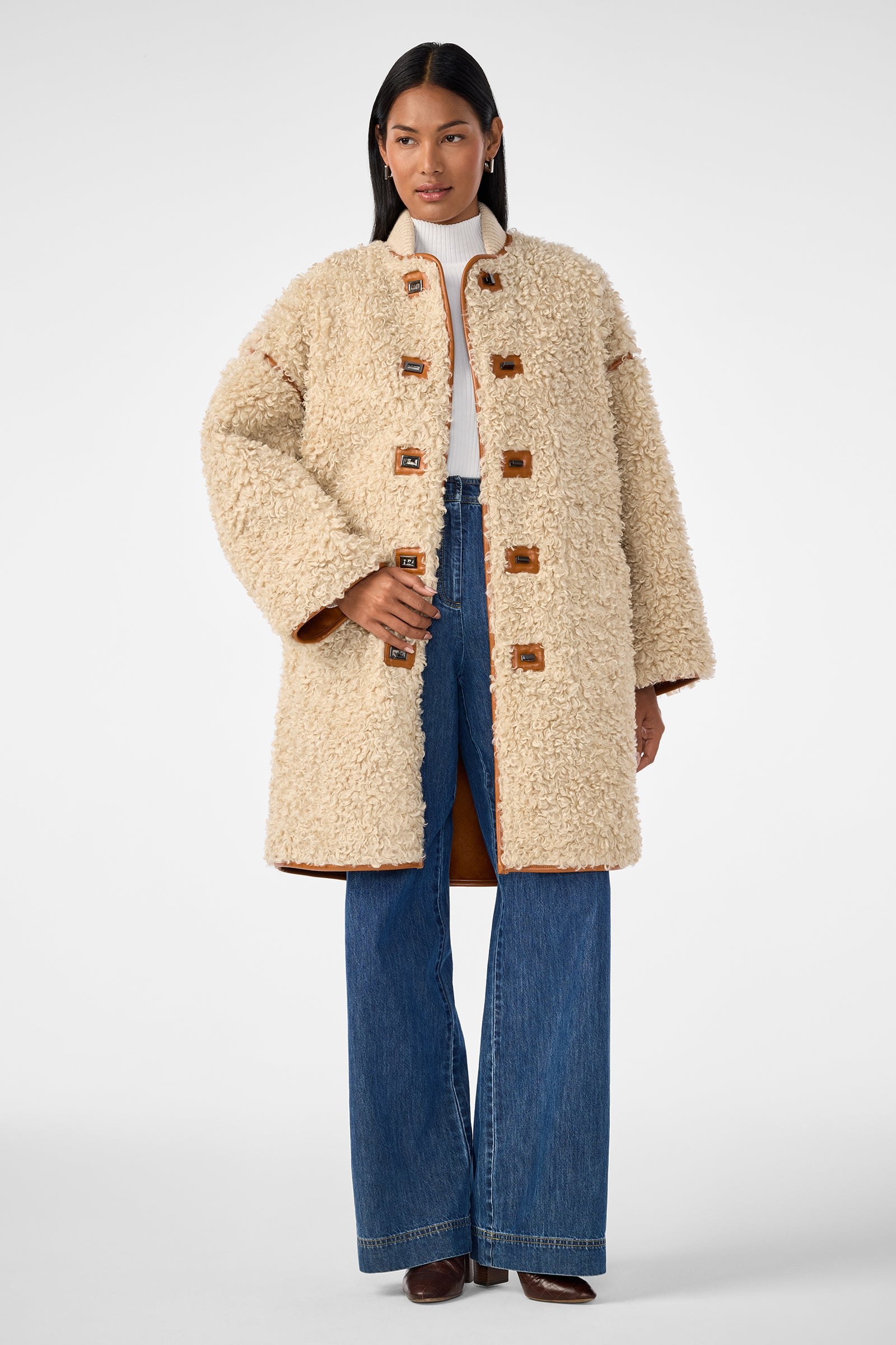 A woman stands against a plain white background wearing the Spartan Coat, a cream-colored textured shearling coat with large toggle buttons, styled with a white turtleneck sweater, wide-leg blue jeans, and brown boots.