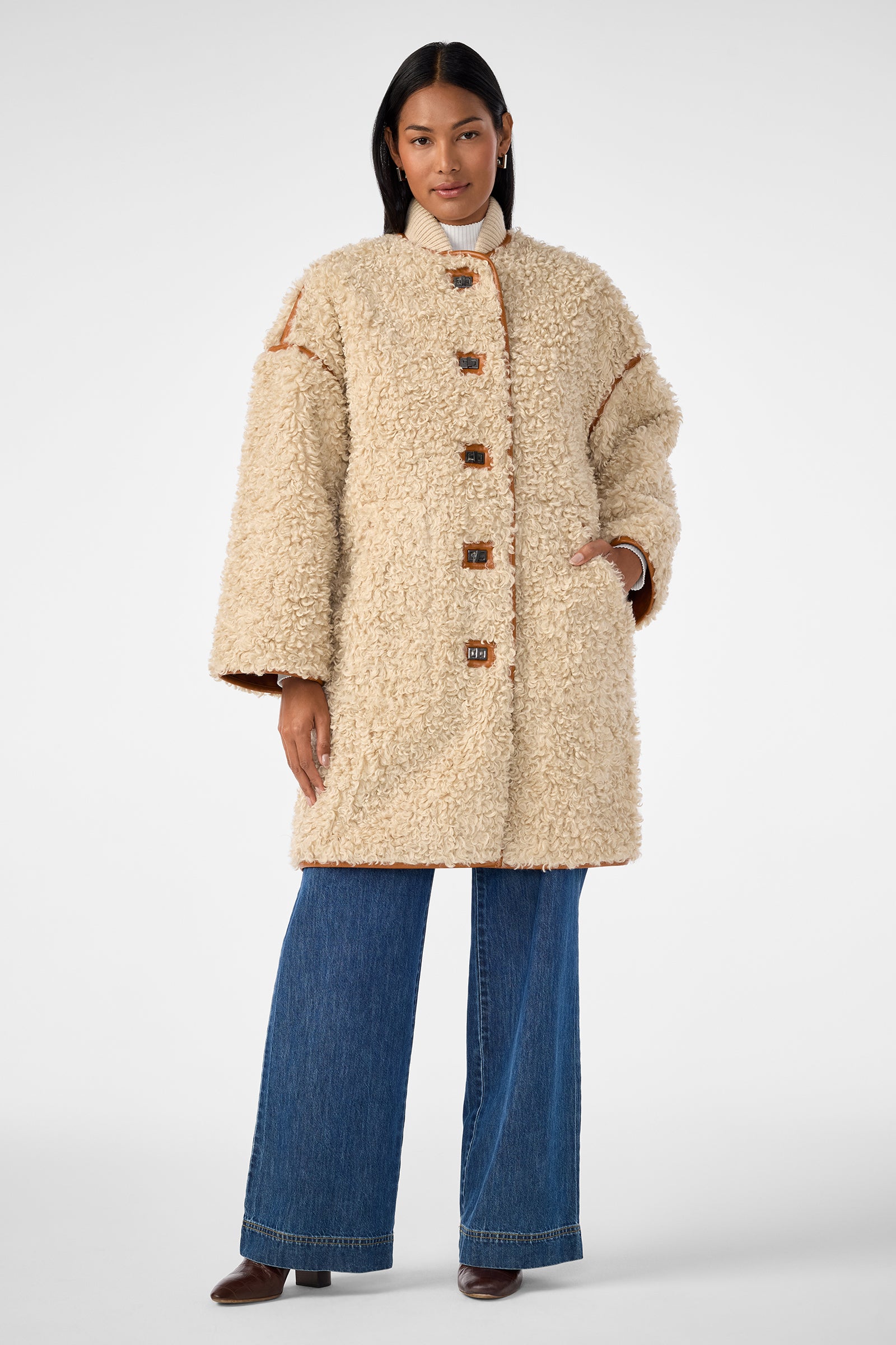 A woman faces forward in a cream Spartan Coat with large buttons, styled with blue wide-leg jeans, a white turtleneck, and brown shoes against a plain background.