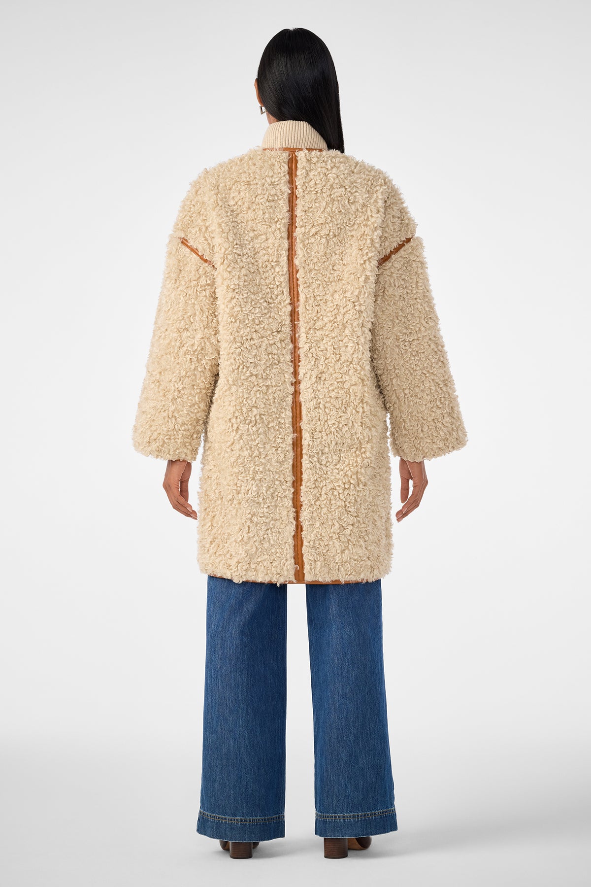 A person with long dark hair, seen from behind, wears the Spartan Coat—a cream-colored, textured shearling coat with tan trim—paired with wide-leg blue jeans and brown shoes against a plain light background.