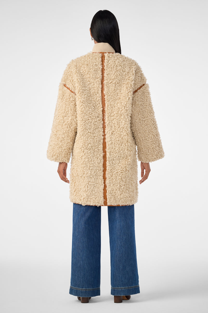 A person with long dark hair, seen from behind, wears the Spartan Coat—a cream-colored, textured shearling coat with tan trim—paired with wide-leg blue jeans and brown shoes against a plain light background.