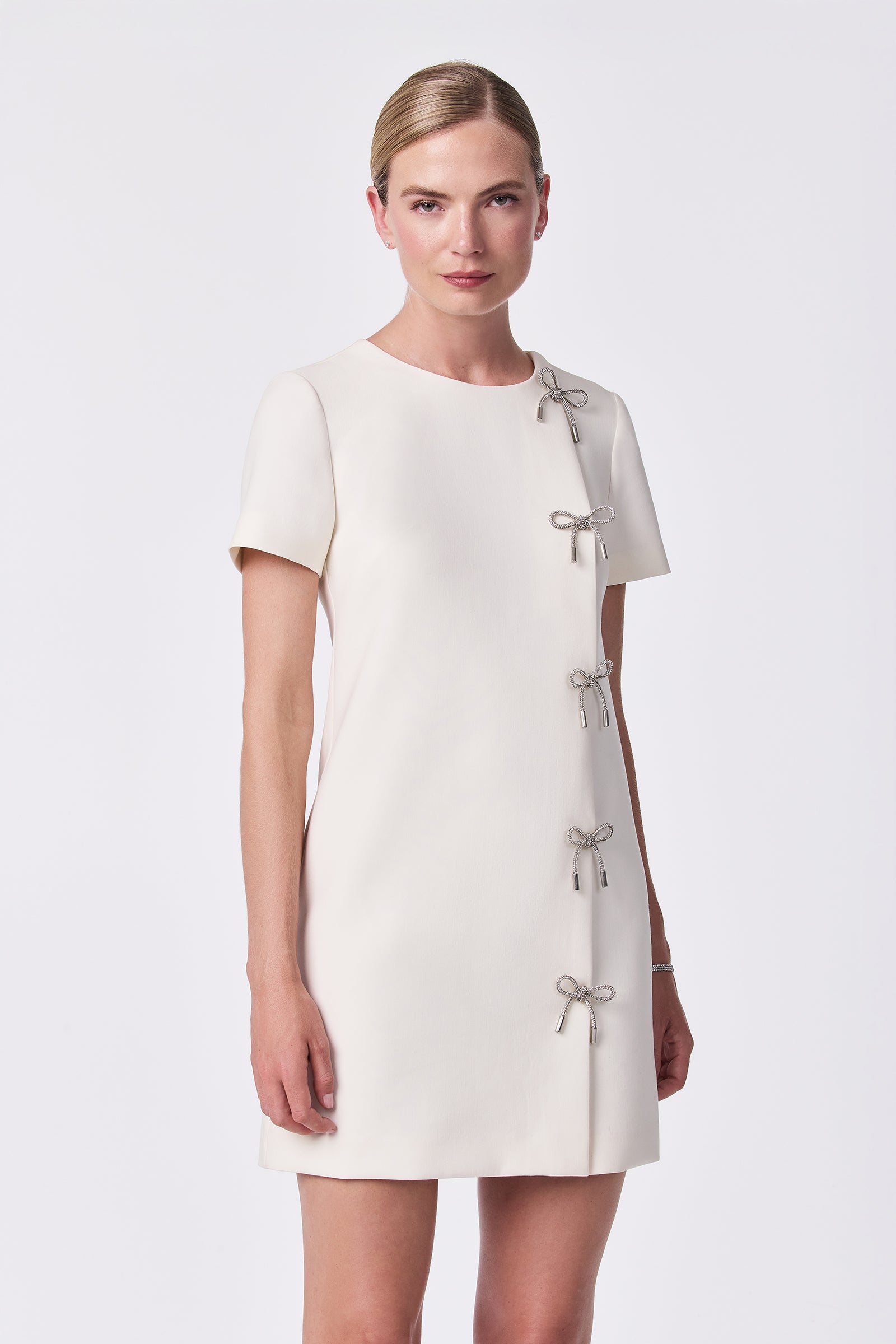 A woman models the Diamonds Crystal Bows Sheath Dress in Winter White, featuring short sleeves and decorative crystal bows along the right side, against a plain white background.