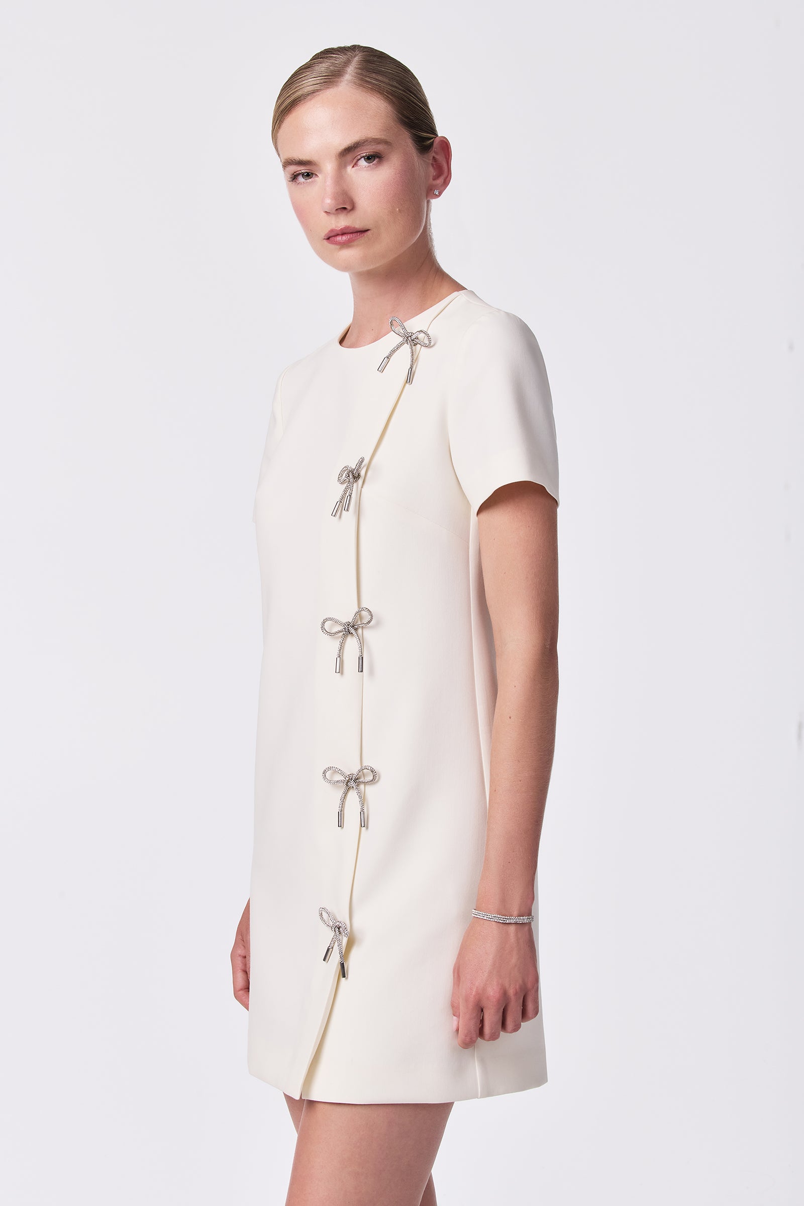 A woman wears the Diamonds Crystal Bows Sheath Dress in Winter White, featuring short sleeves and silver bow accents along a diagonal seam, standing against a plain white background.