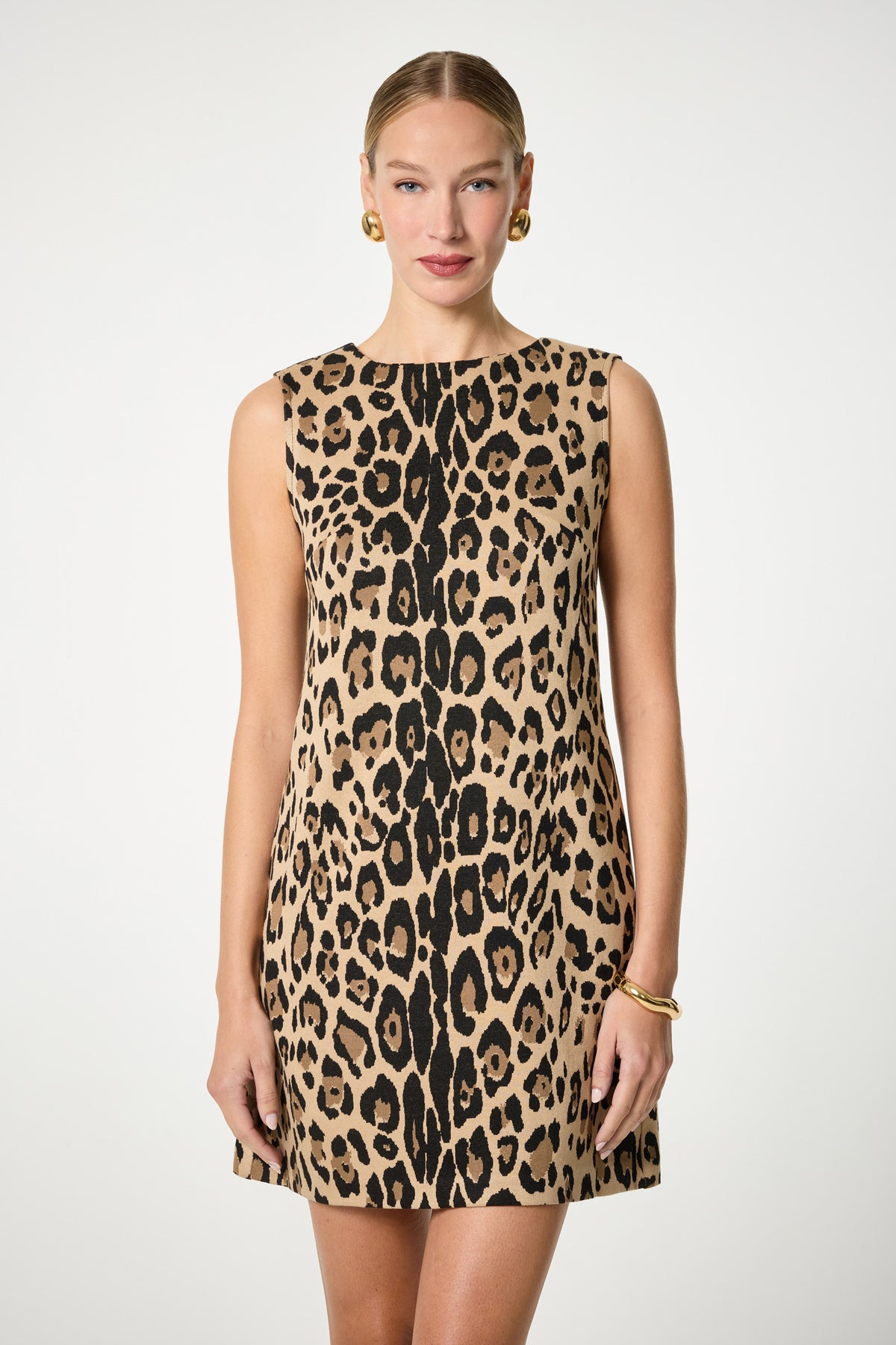 A woman faces forward wearing the Adapted Sleeveless Shift Dress, featuring a bold leopard print and knee-length cut. Her hair is pulled back, and she accessorizes with gold hoop earrings and a bracelet against a plain white background.