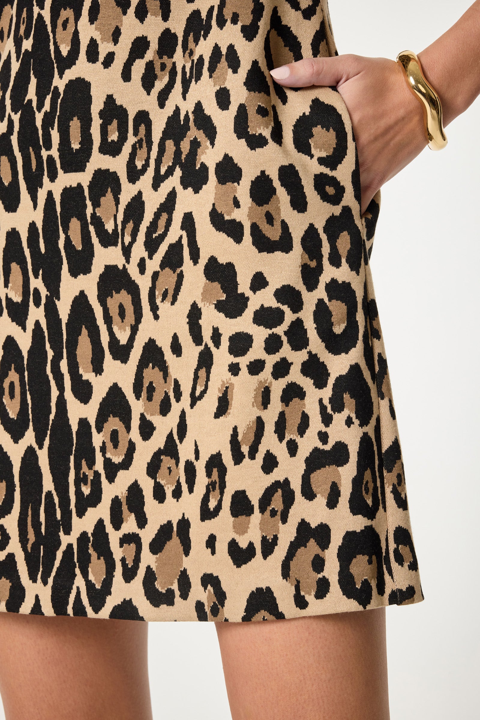 A close-up of a person wearing the Adapted Sleeveless Shift Dress in tan and black leopard print with one hand in the pocket, accessorized with a chunky gold bracelet.