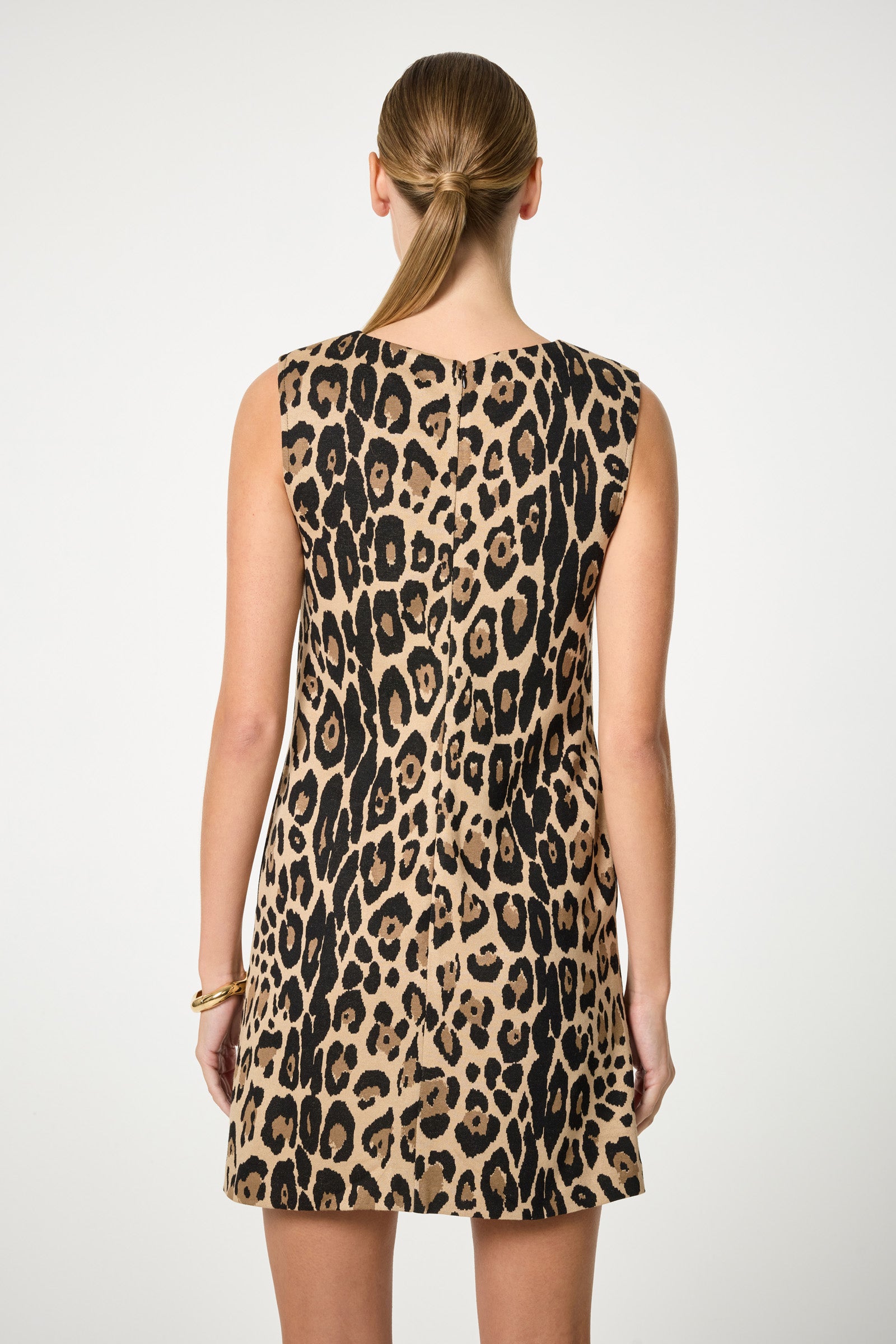 A person with straight light brown hair in a low ponytail is wearing the Adapted Sleeveless Shift Dress, a knee-length dress with a bold leopard print, viewed from the back against a plain white background.