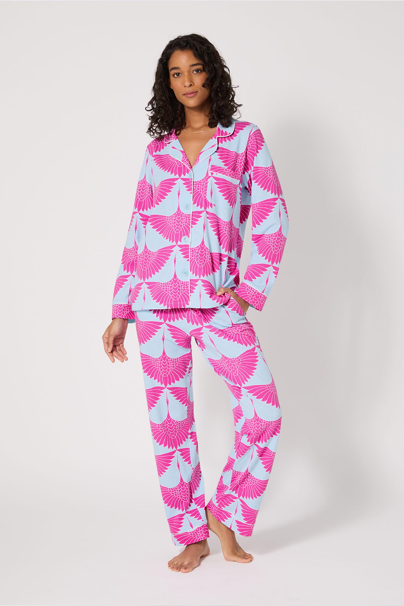 A woman stands barefoot in the Classic PJ Set - Cranes, featuring matching light blue pajamas with a bold pink crane pattern, button-up shirt, and long pants against a plain white background.