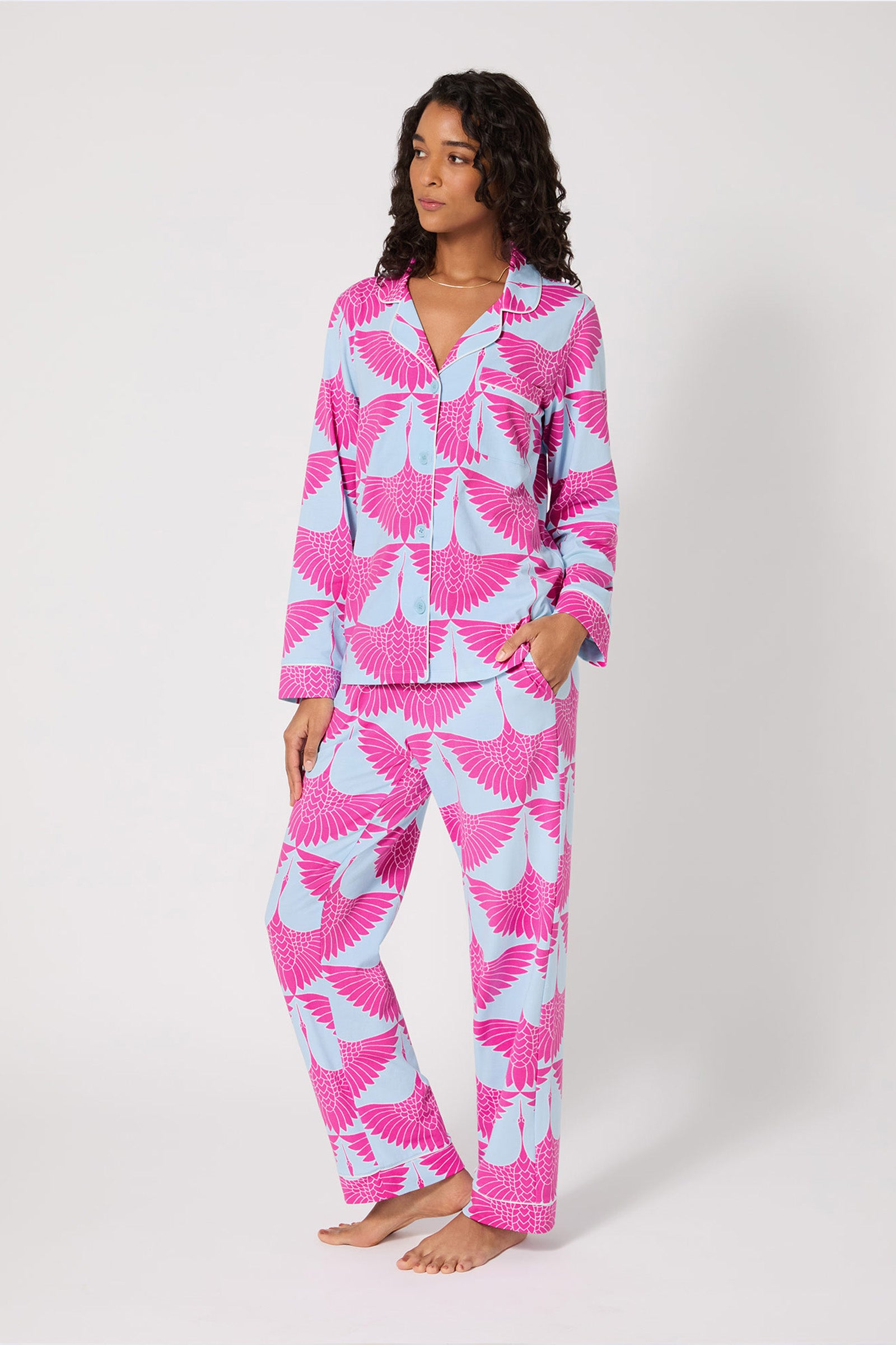 A woman stands barefoot against a plain backdrop, wearing the Classic PJ Set - Cranes, featuring a bold pink and blue abstract bird print with long sleeves, button-down top, and straight-leg pants.