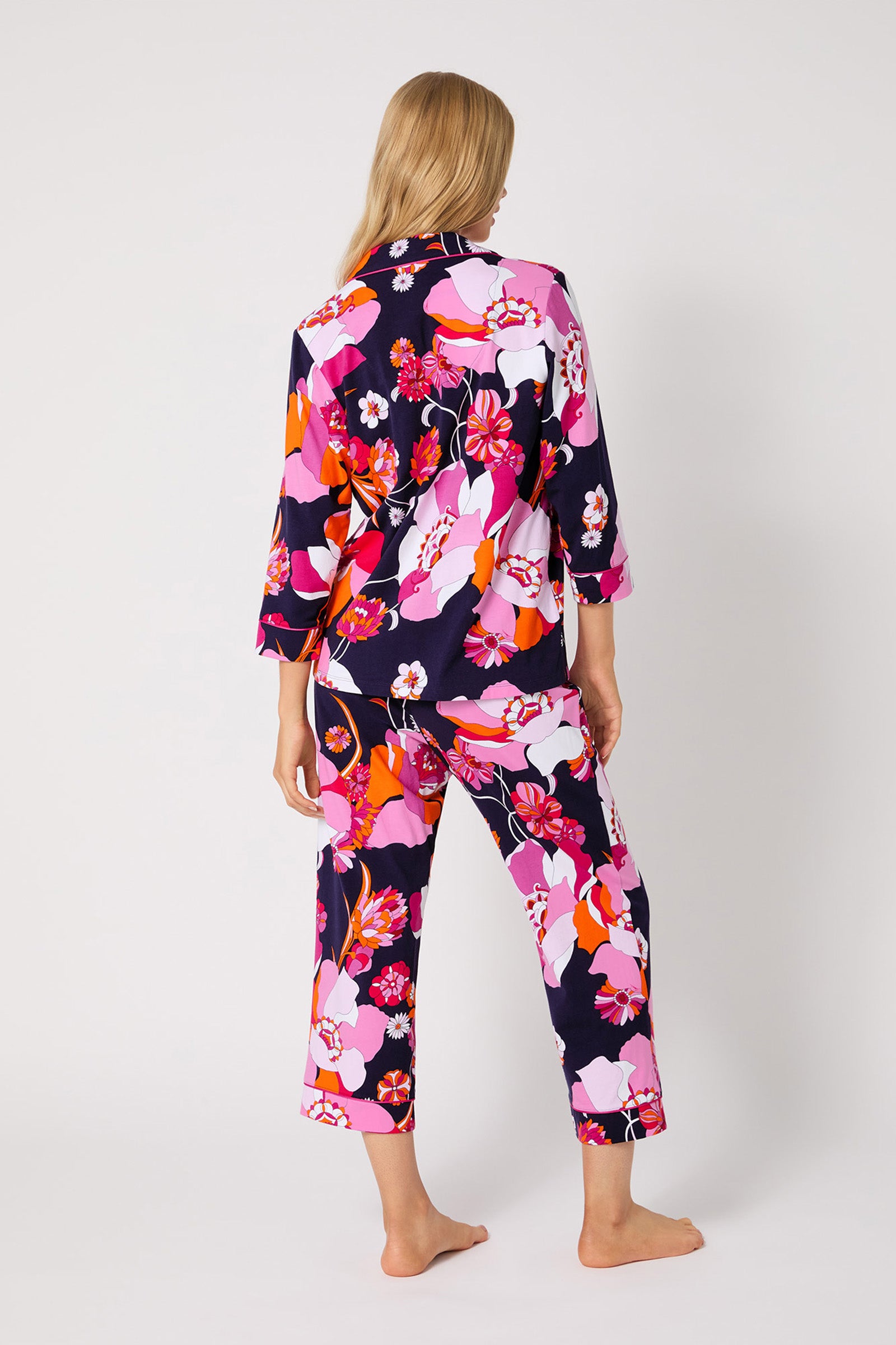 A woman stands barefoot facing away, wearing the Crop Classic PJ Set - Floral Medley, featuring a vibrant floral pattern in pink, orange, white, and purple on a dark background.