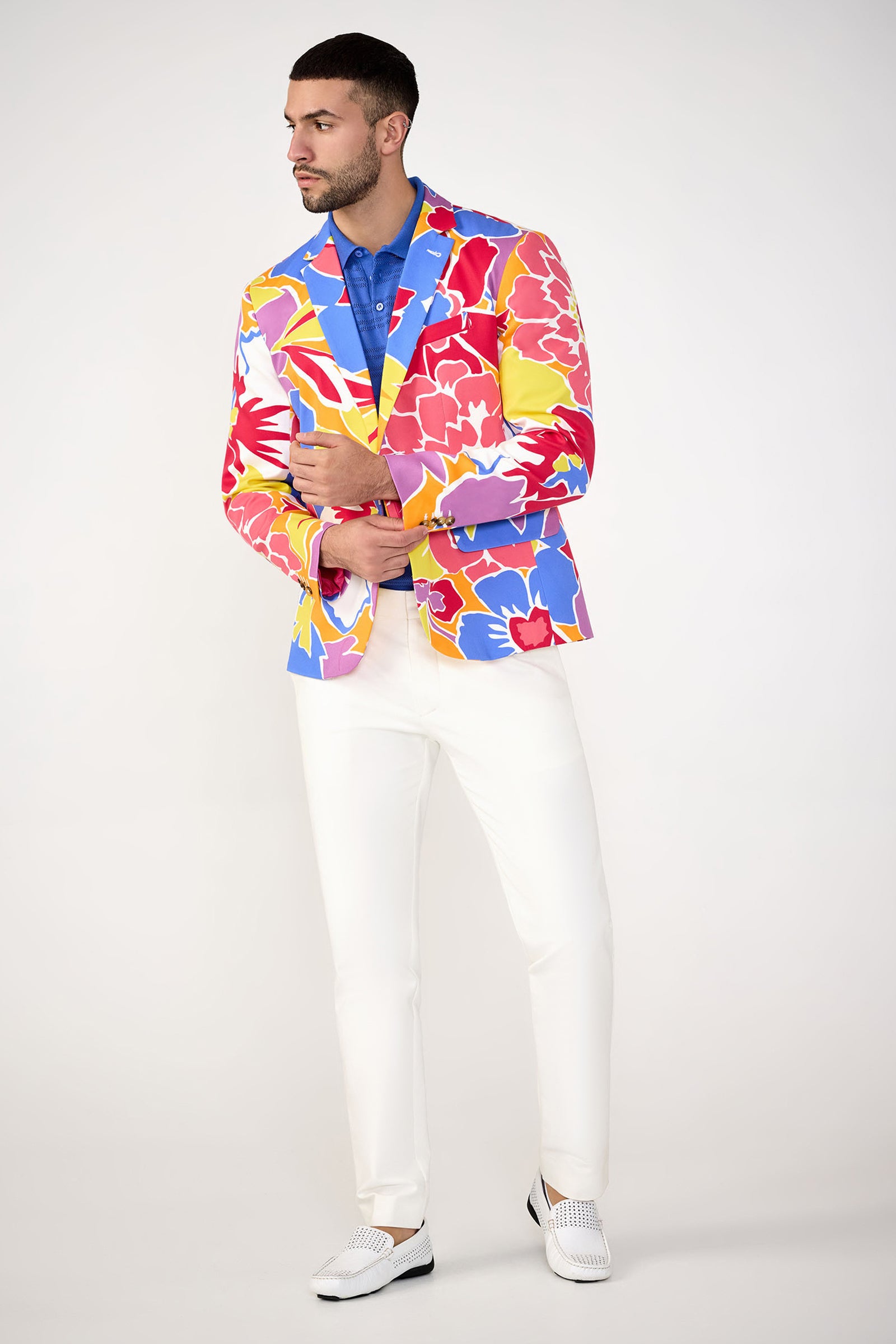 A man wears the MR TURK Thurston Blazer - Soie Paradis, featuring a vibrant floral print, over a blue shirt, white pants, and white shoes against a plain light background.