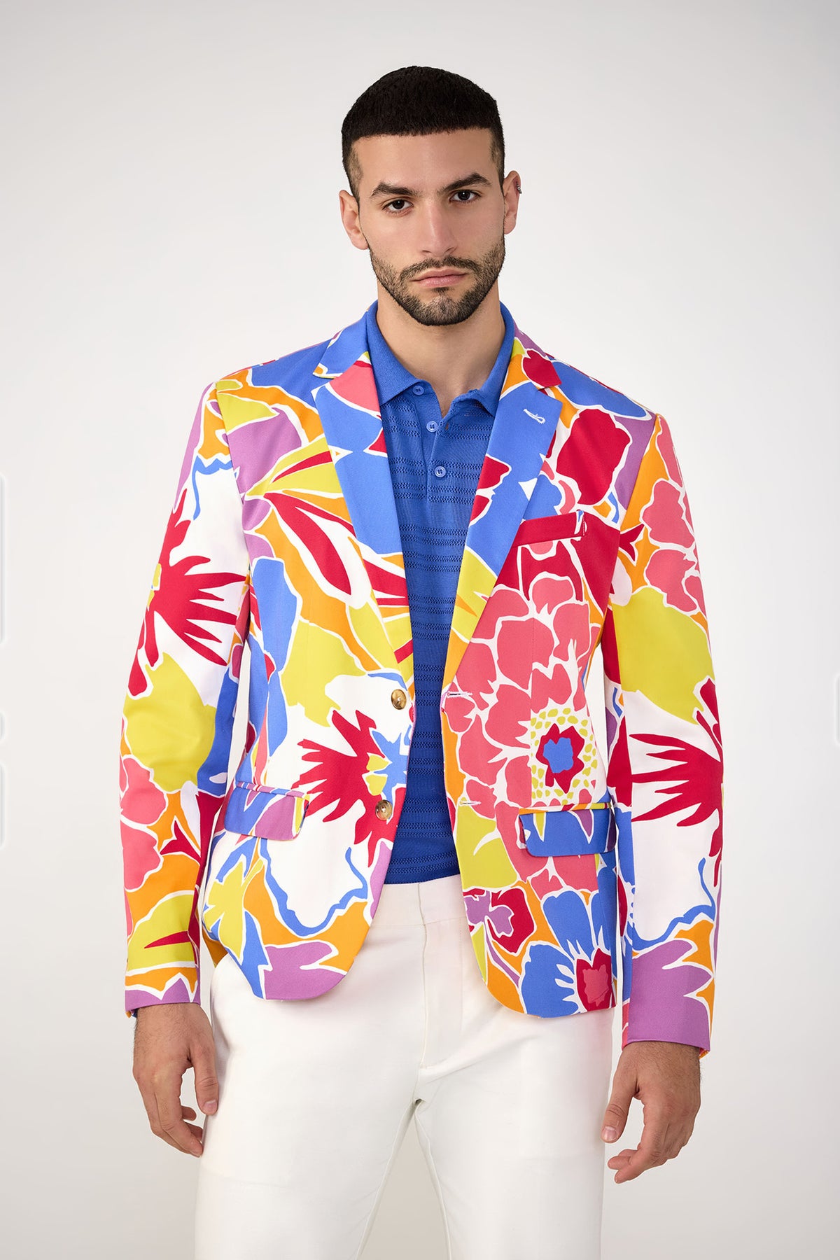 A man wears the Thurston Blazer - Soie Paradis by MR TURK over a blue polo and white pants, standing against a plain light background and looking directly at the camera with a neutral expression.