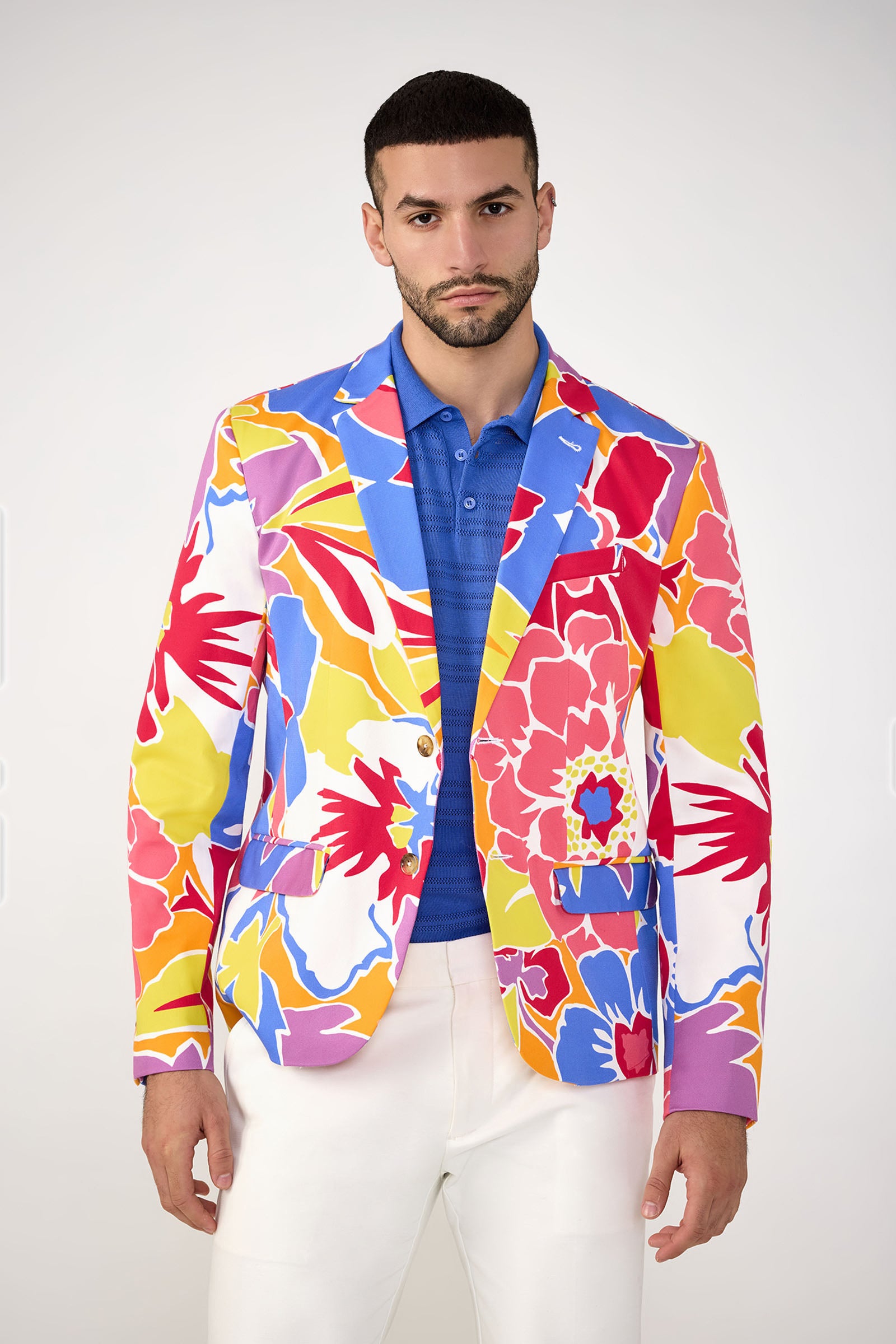 A man wears the Thurston Blazer - Soie Paradis by MR TURK over a blue polo and white pants, standing against a plain light background and looking directly at the camera with a neutral expression.