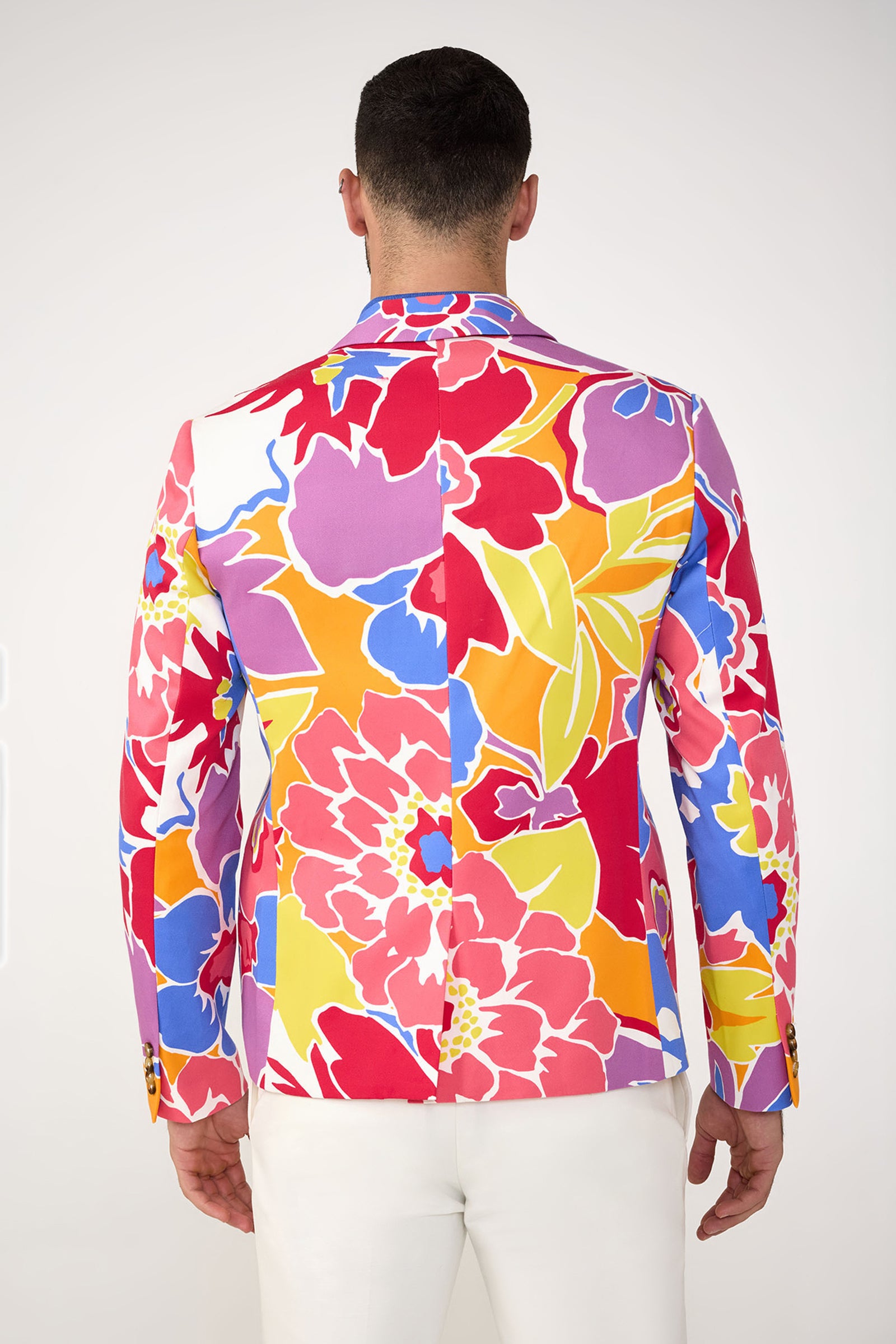 A person stands facing away in the MR TURK Thurston Blazer - Soie Paradis, featuring a bold, colorful floral print in red, pink, yellow, purple, and blue, paired with white pants against a plain background.