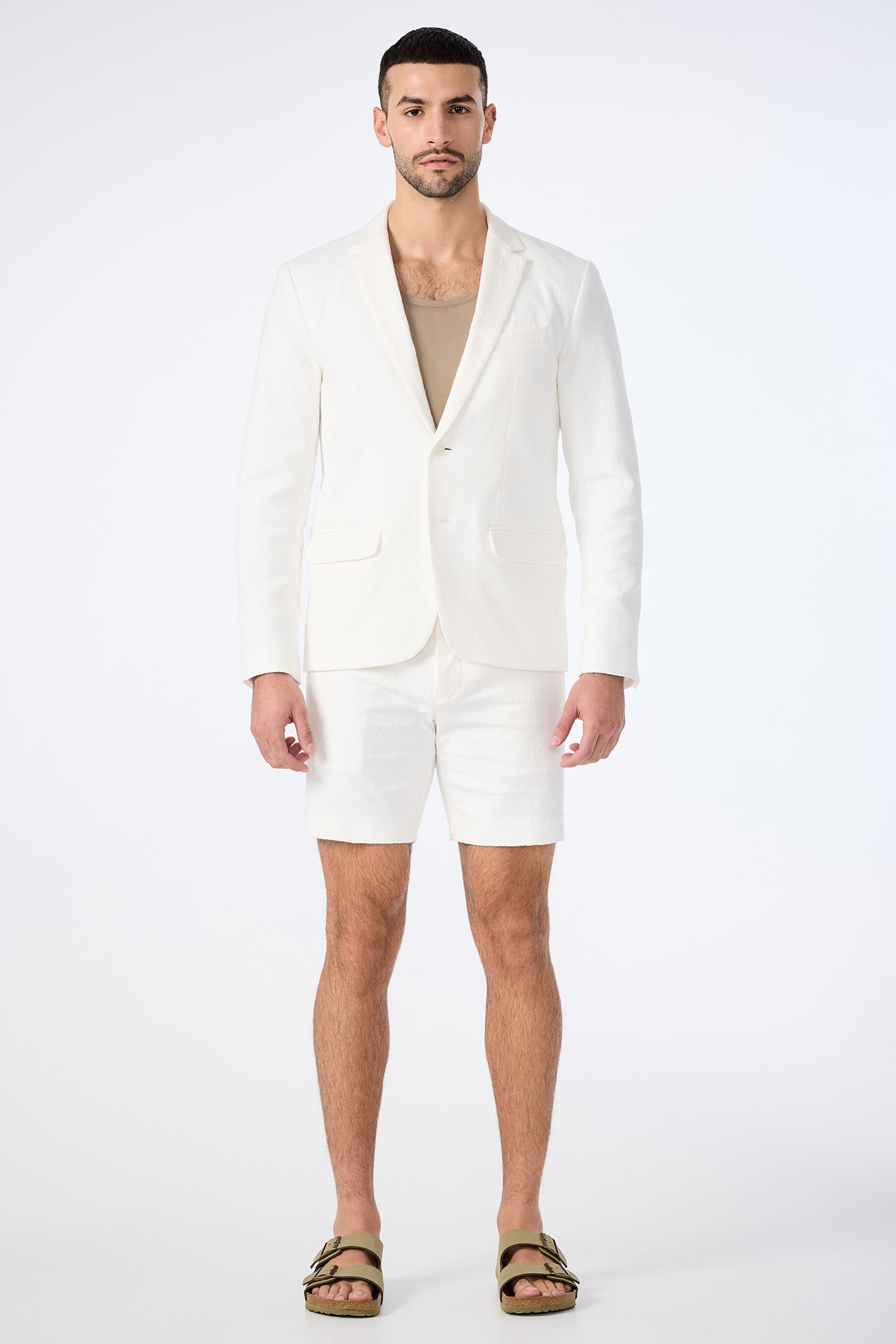 A man faces forward against a light background, wearing the Thurston Blazer - Bi-Stretch Cotton in white with matching shorts, a beige shirt underneath, and olive green sandals.