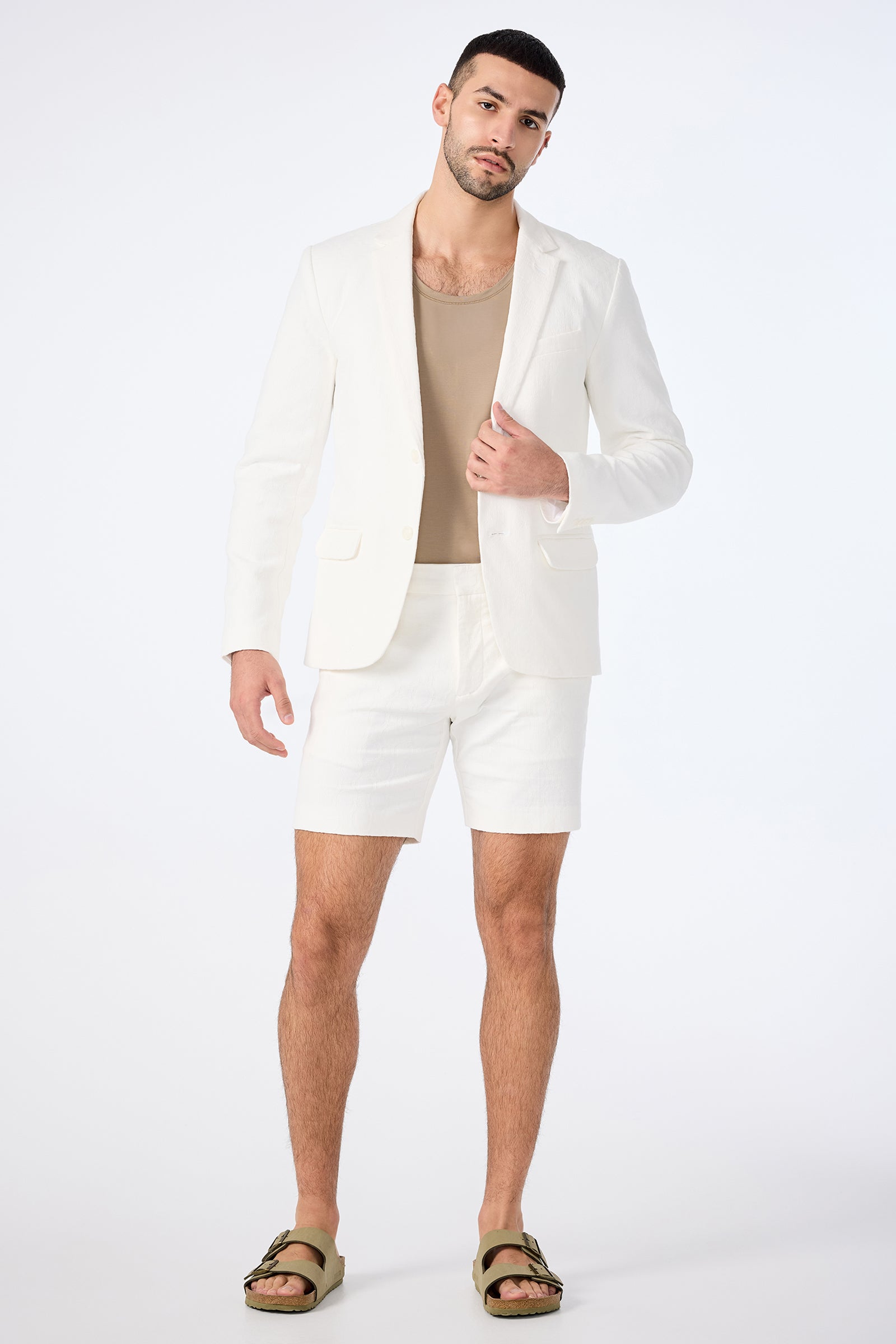 A man stands against a plain white background, looking relaxed at the camera while wearing the Thurston Blazer - Bi-Stretch Cotton in white, with matching shorts, a beige tank top, and green sandals.