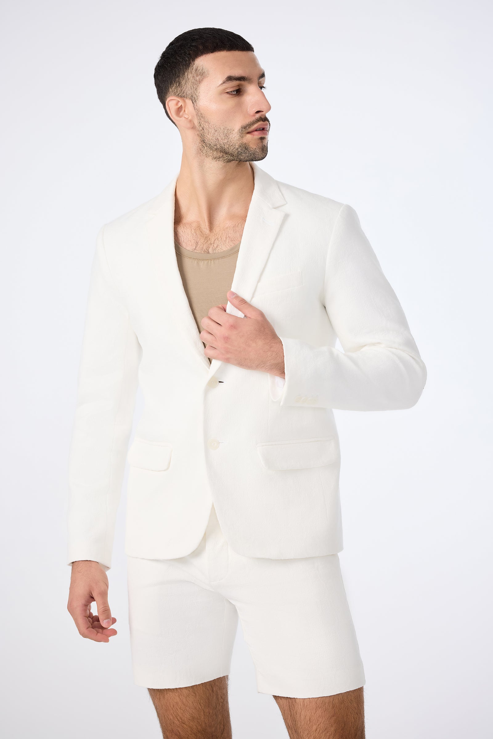 A man in the Thurston Blazer - Bi-Stretch Cotton in Whitewash, paired with matching shorts and a beige shirt, poses against a plain white background, glancing to the side while adjusting his jacket.