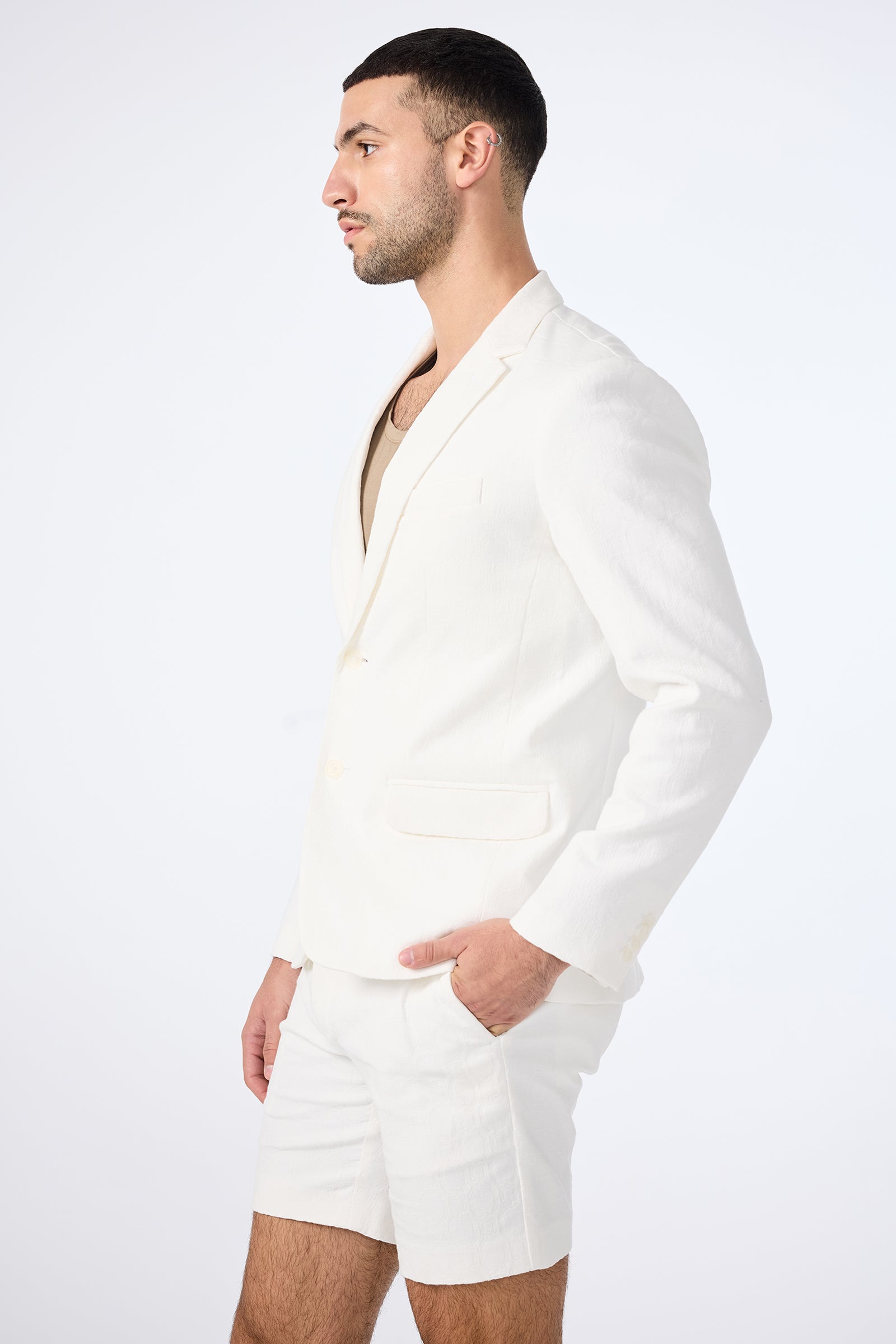 A man in the Thurston Blazer - Bi-Stretch Cotton and matching white shorts stands in profile against a plain light background, his left hand in his pocket and short dark hair neatly styled.