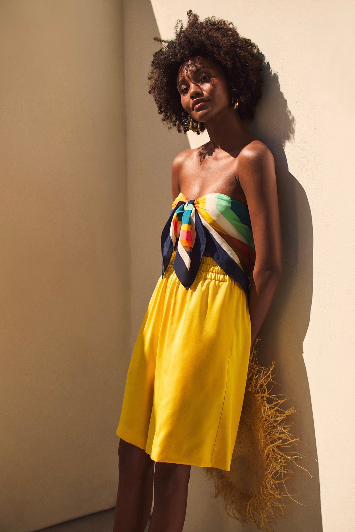 A woman with curly hair leans against a beige wall, wearing a strapless colorful striped top and the Chiana Satin Shorts, holding a straw hat with fringe as sunlight creates soft shadows on her and the wall.