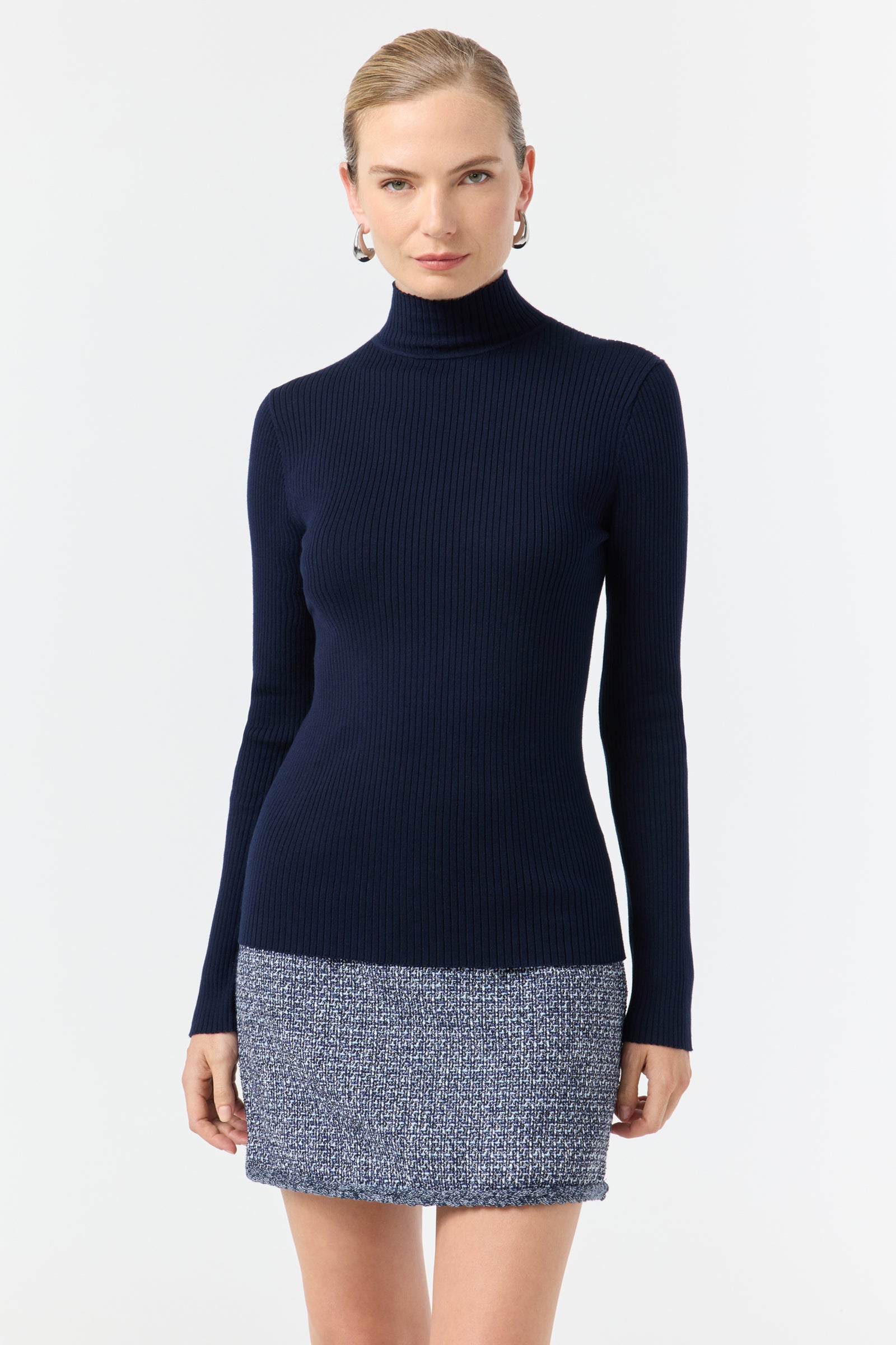 A woman wears the Hempstead Turtleneck Sweater in Indigo, a fitted, ribbed style, paired with a short blue and white textured skirt, standing against a plain white background.