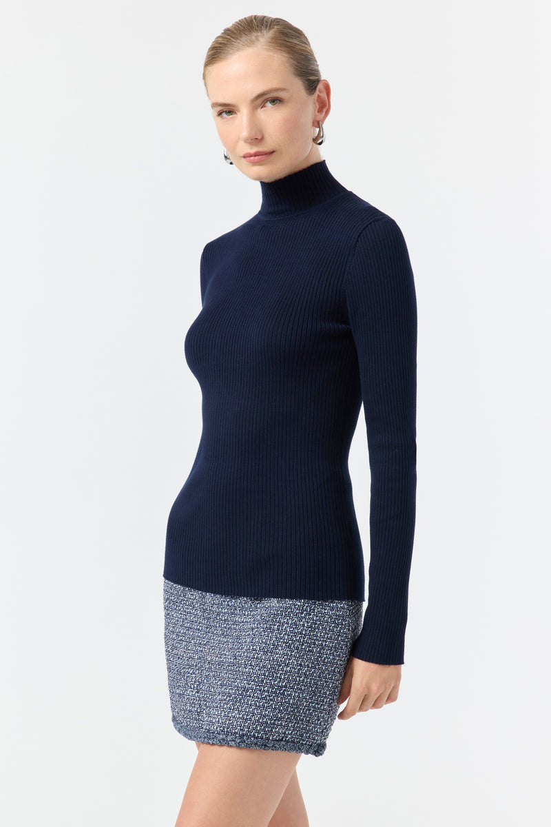 A woman with blonde hair in a sleek bun wears the fitted Hempstead Turtleneck Sweater in Indigo, paired with a short blue skirt, standing confidently against a plain white background.