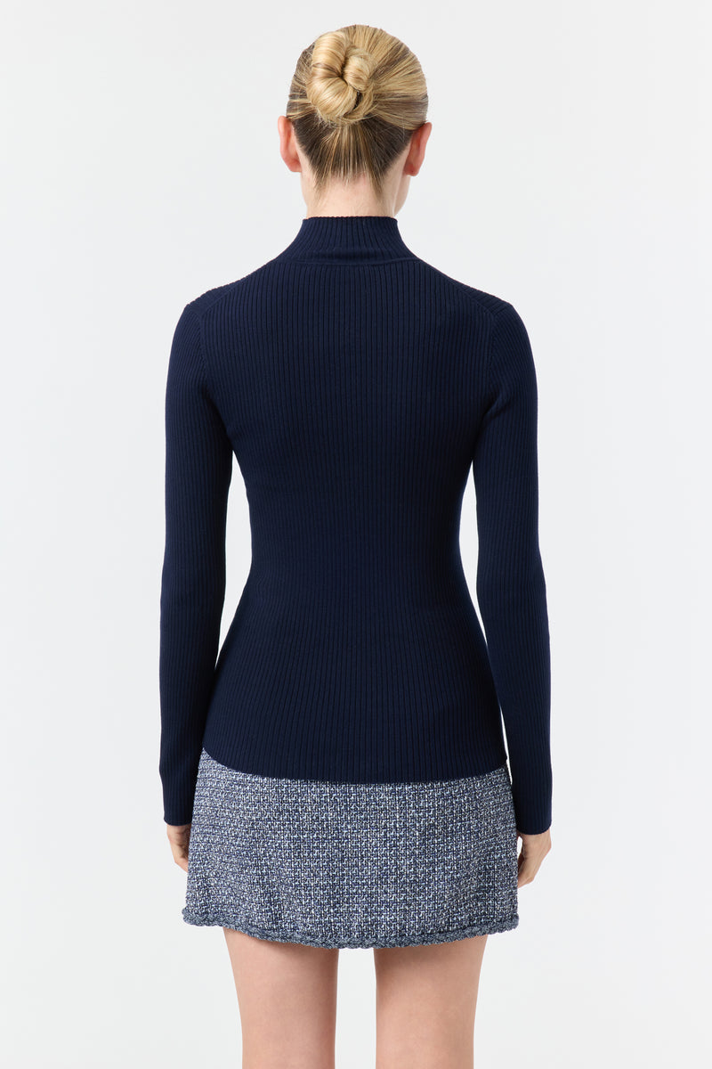 A woman with blond hair in a bun faces away, wearing the Hempstead Turtleneck Sweater in Indigo. The background is plain white.