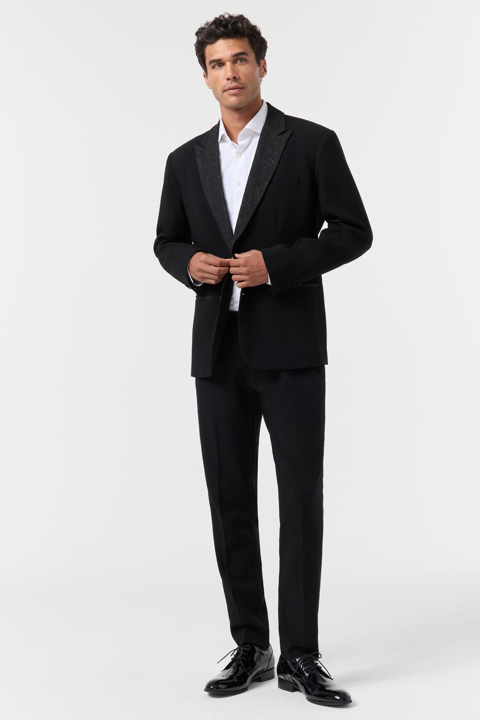 A man stands against a white background, wearing the Miles Blazer over a white dress shirt and black dress shoes. He is buttoning the jacket and gazes slightly upward with a neutral expression.