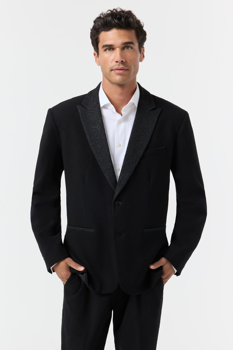 A man with dark hair models the Miles Blazer—black suit jacket and white dress shirt—standing forward with hands in his pockets against a plain white background.