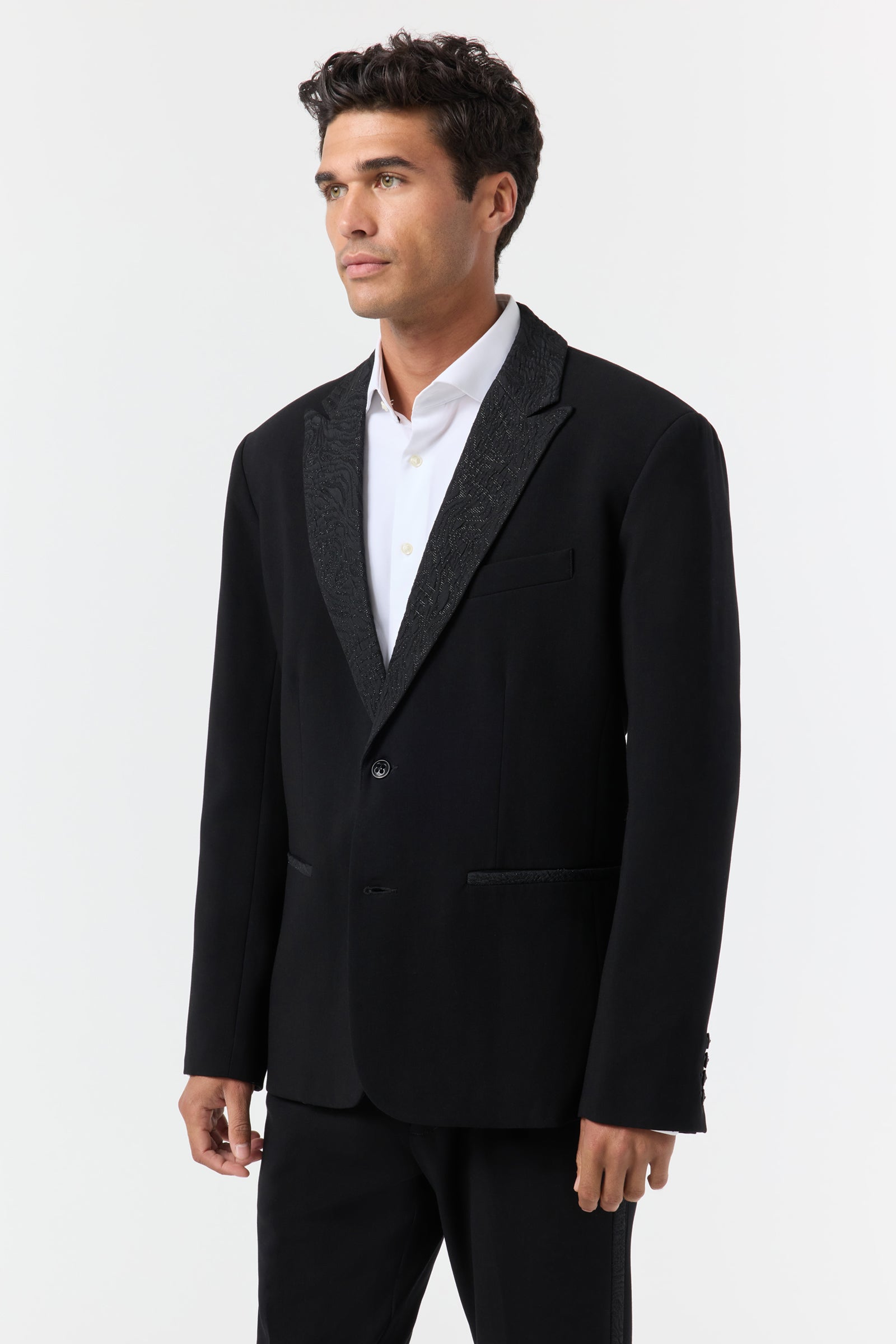 A man stands facing slightly left, wearing the Miles Blazer—a black suit jacket with a textured lapel—over a white dress shirt. The background is plain and light-colored.