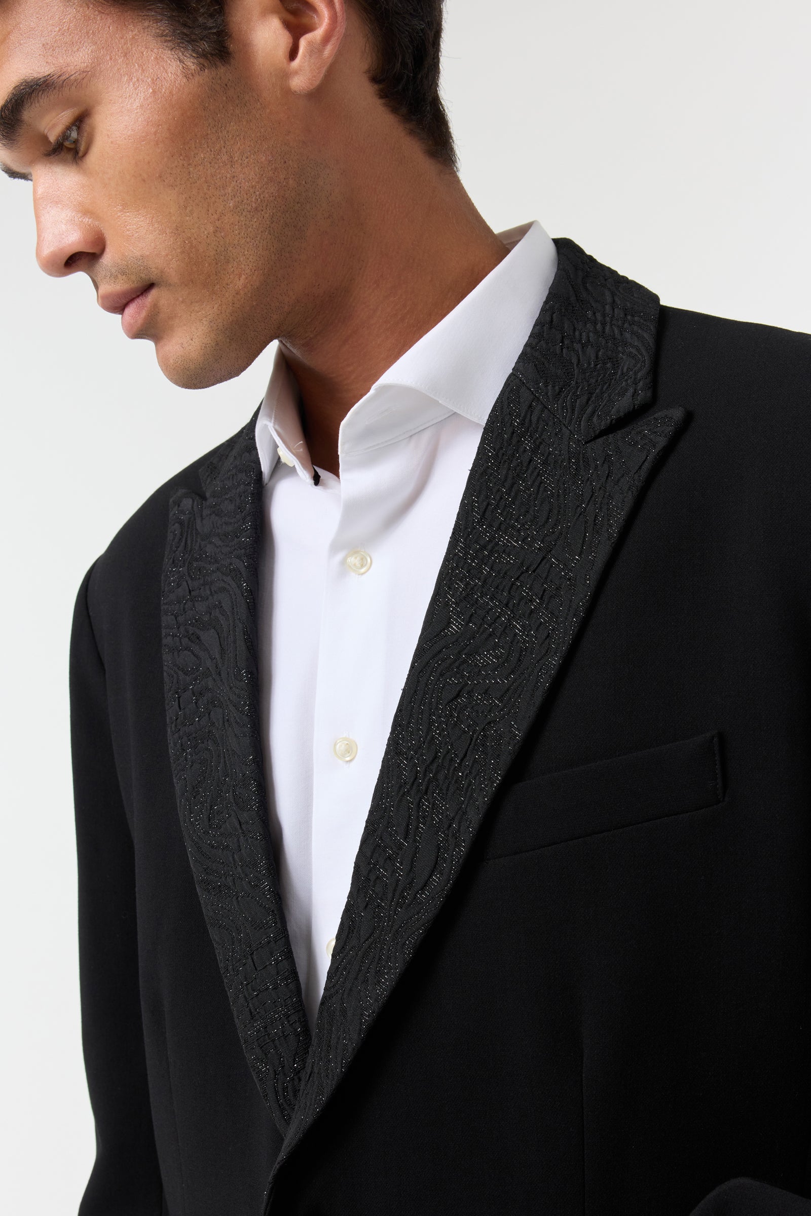 A man wears the Miles Blazer—a black tuxedo jacket with a textured, patterned lapel—over a white dress shirt, looking down and away from the camera against a plain background.