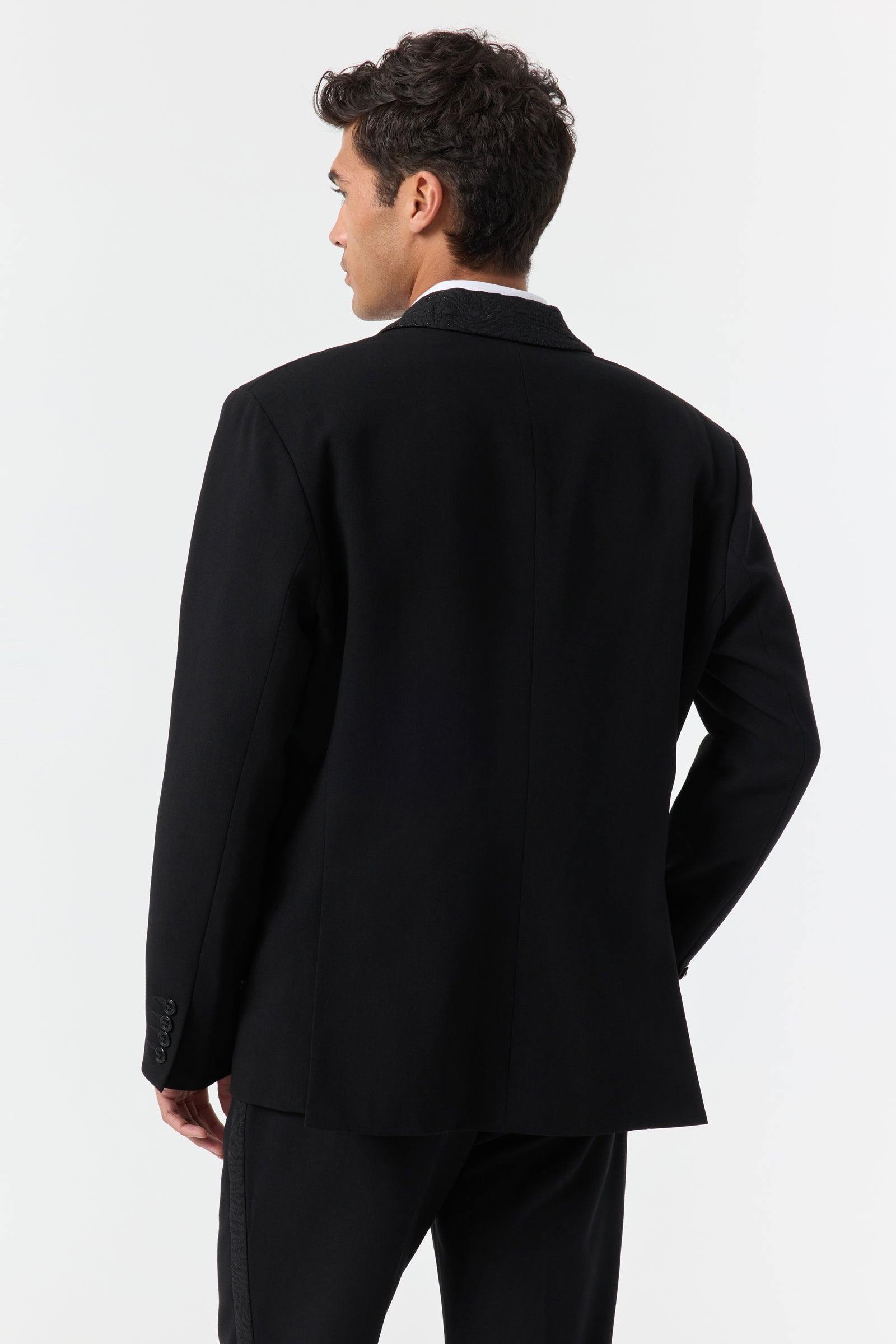 A man with short, dark, curly hair stands facing away against a plain white background, wearing the Miles Blazer and matching black pants, with his left hand resting on his hip.