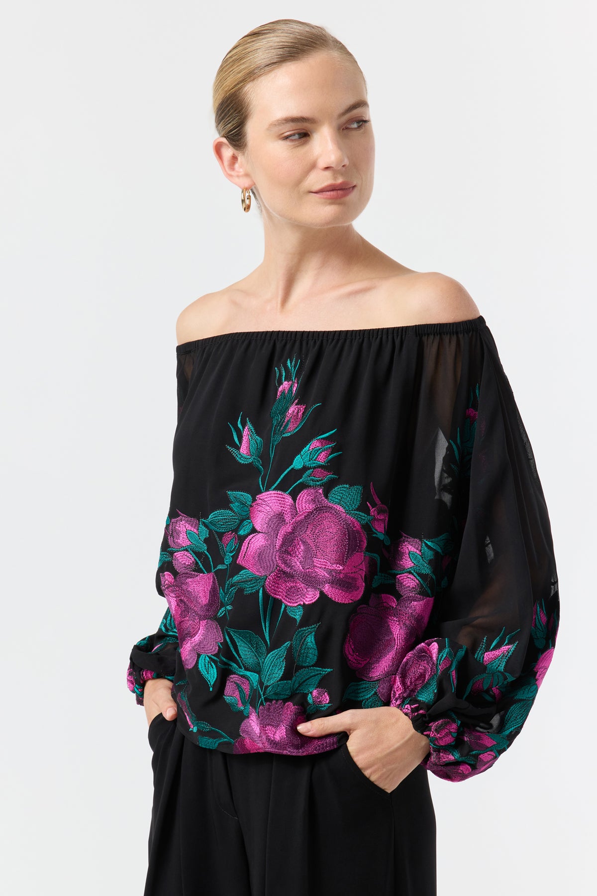 Woman wearing the Soho 2 Embroidered Top—an off-the-shoulder black blouse with pink and green floral embroidery and long sleeves—paired with black pants. Her light hair is pulled back as she smiles softly, looking to the side.