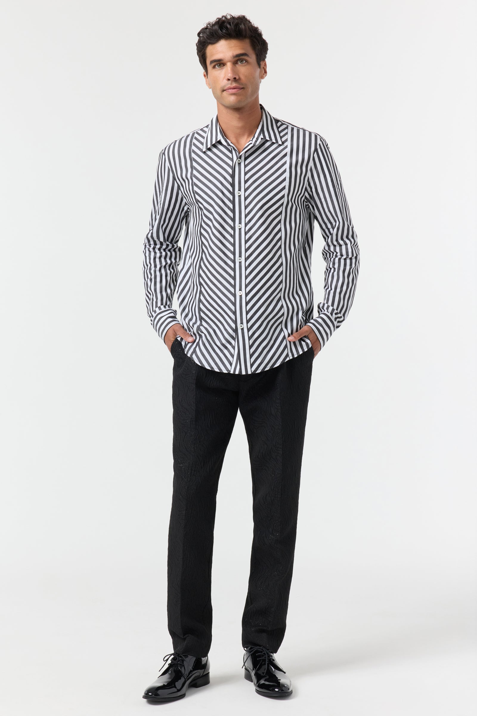 A man stands against a plain white background wearing the Brody Tux Shirt, which features black and white vertical stripes, paired with black pants and dress shoes. His hands rest in his pockets as he looks slightly upwards.