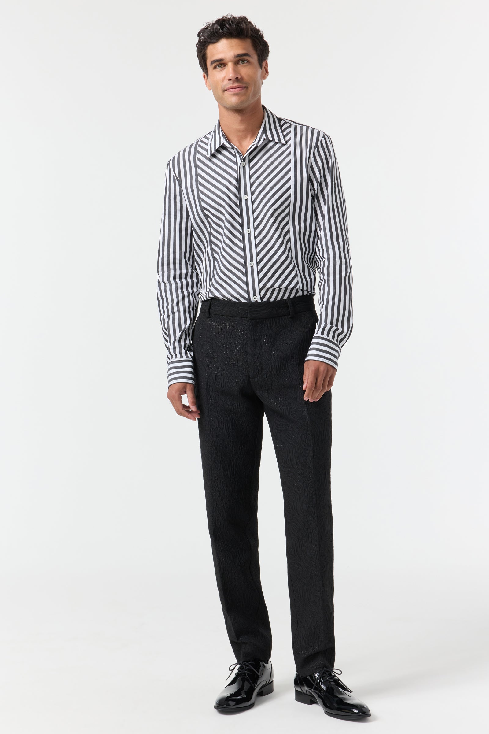 A man stands against a plain white background wearing a black and white vertical striped shirt, Clyde Slim Trouser - Metallic Groove, and black dress shoes, with a neutral expression.