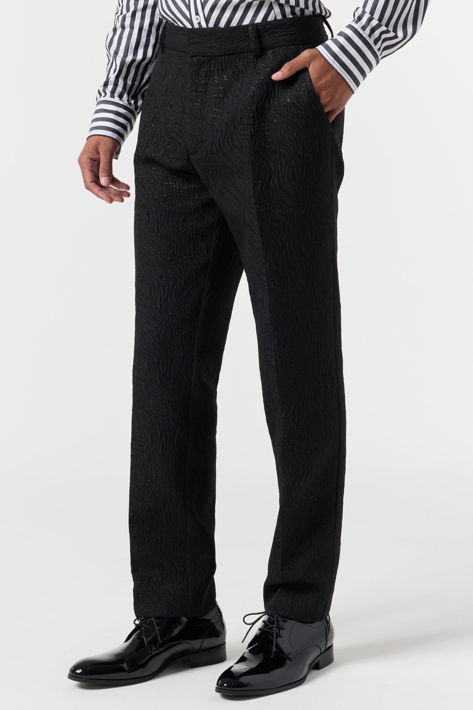 A person wears the Clyde Slim Trouser - Metallic Groove, styled with a black and white striped dress shirt and shiny black dress shoes. Only the lower half is visible against a plain white background.