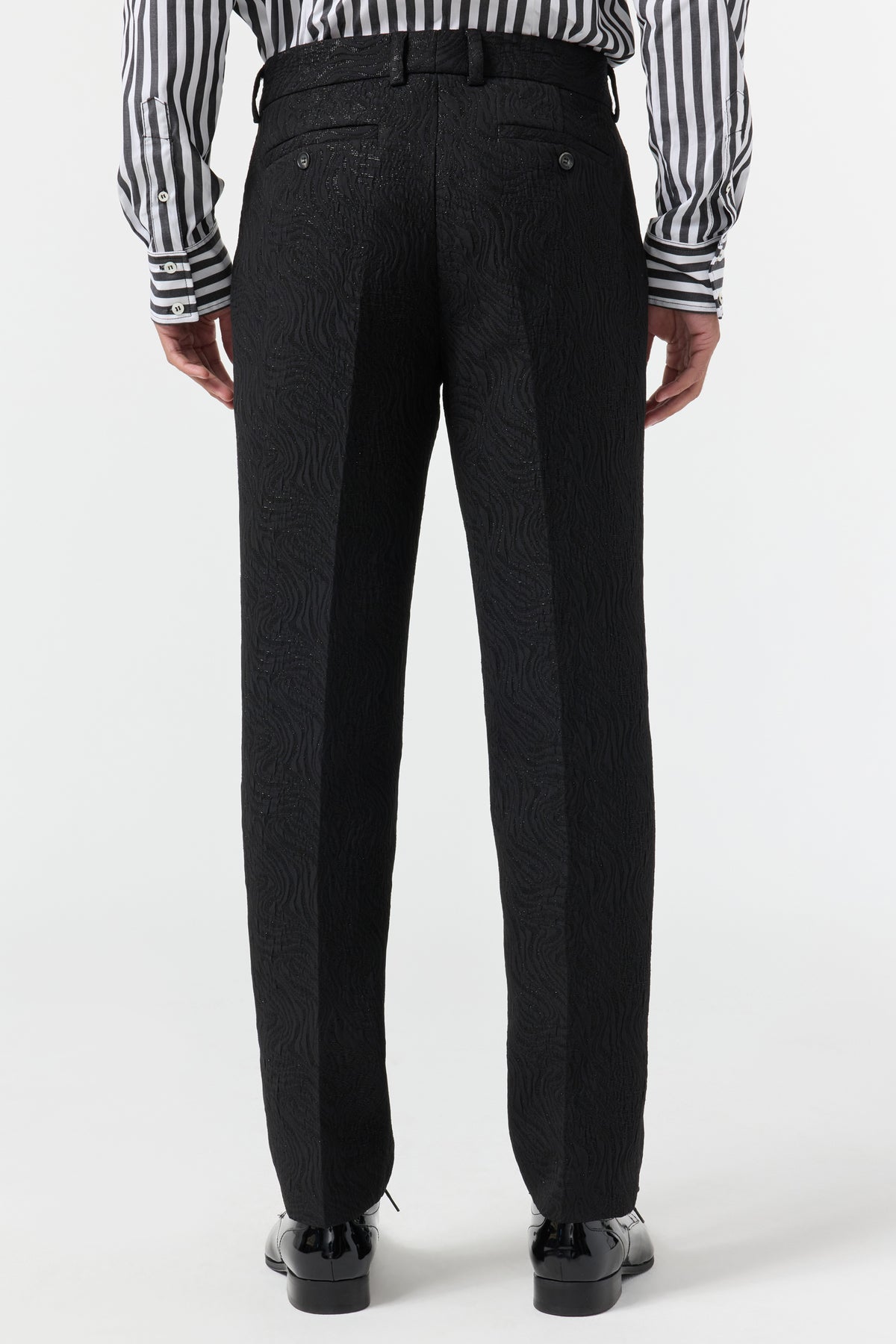 A person faces away, wearing Clyde Slim Trouser - Metallic Groove, a black belt, shiny black shoes, and a black-and-white striped long-sleeve shirt against a plain white background.